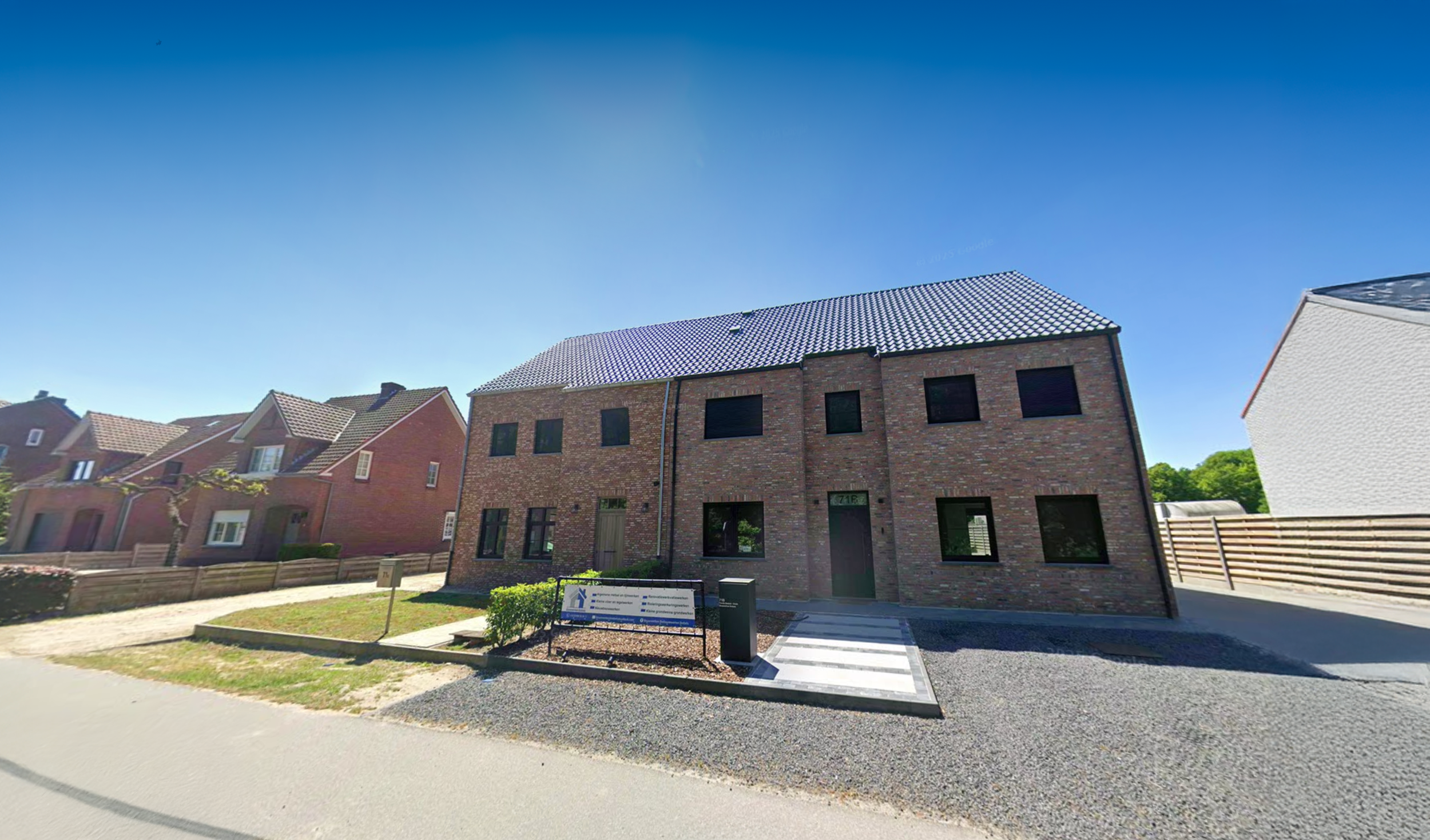 Nieuwe moderne stenen woning met zwarte ramen en een deur, kleine tuin met struik en bank, witte stoep en een stalen hek, onder een heldere blauwe lucht.