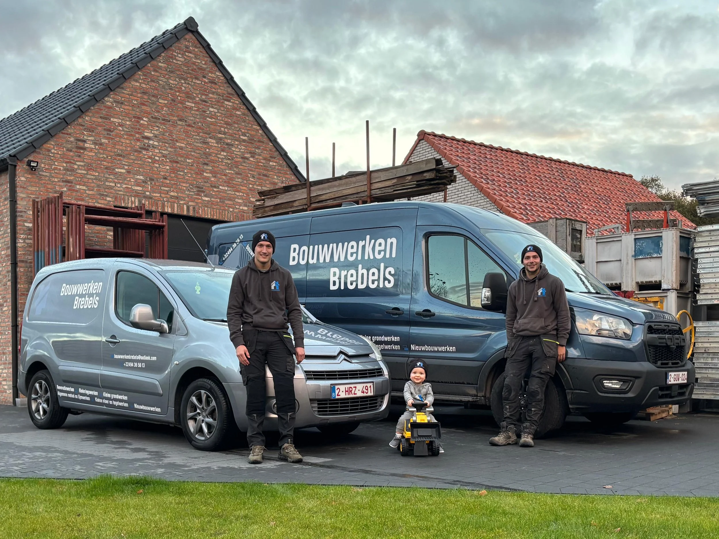 Twee mannen en een kind staan voor twee bedrijfsvoertuigen die behoren tot Bouwwerken Brebels. De man en het kind lachen en de man heeft zijn hand in zijn pocket. De voertuigen staan op een oprit voor een huis onder een bewolkte sky.