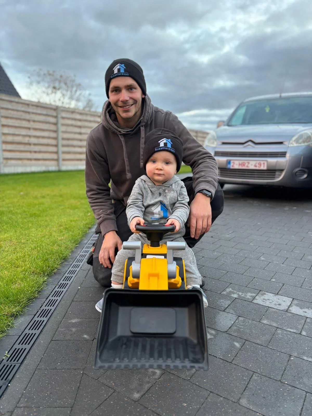 Een man en een kind, beiden gekleed in grijze en zwarte kleding met bouwbedrijf logo, poseren buiten op een oprit met een speelgoedweggebruikerswagen. Een auto staat op de achtergrond. De man is knielend naast het kind, dat op de speelgoedauto zit en naar de camera kijkt. Het weer is bewolkt.