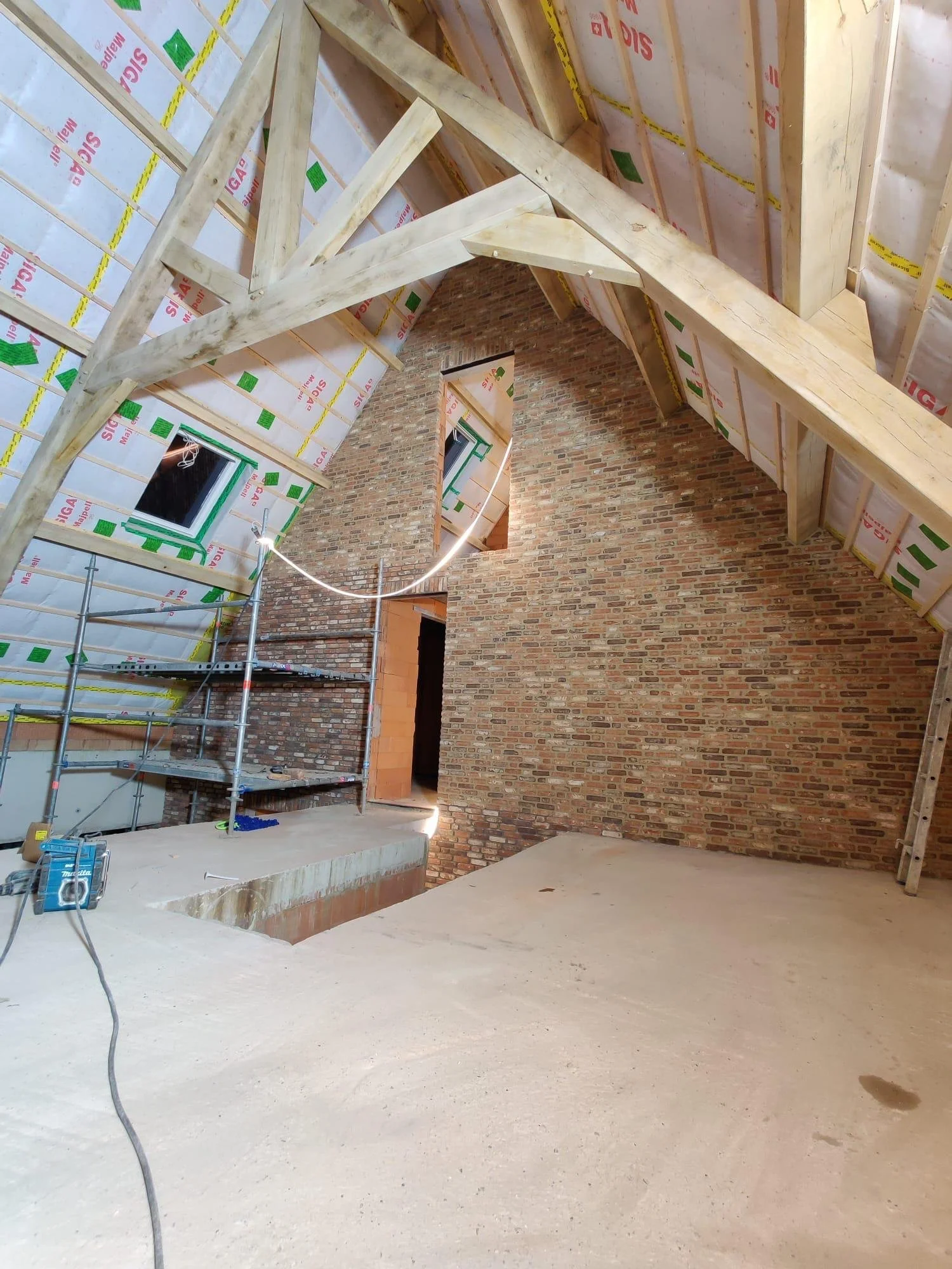 Een binnenkant van een zolderruimte onder constructie, met ruwe vloer, bakstenen muur, houten dakspanten en geïnstalleerde ramen.