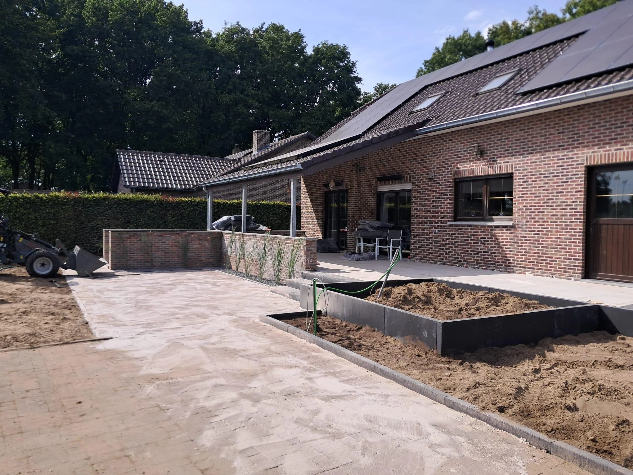 Tuinrenovatie met bestrating, plantenbakken, en voorbereidingen voor nieuwe tuinbedden voor een huis met een bakstenen gevel en zonnepanelen op het dak.