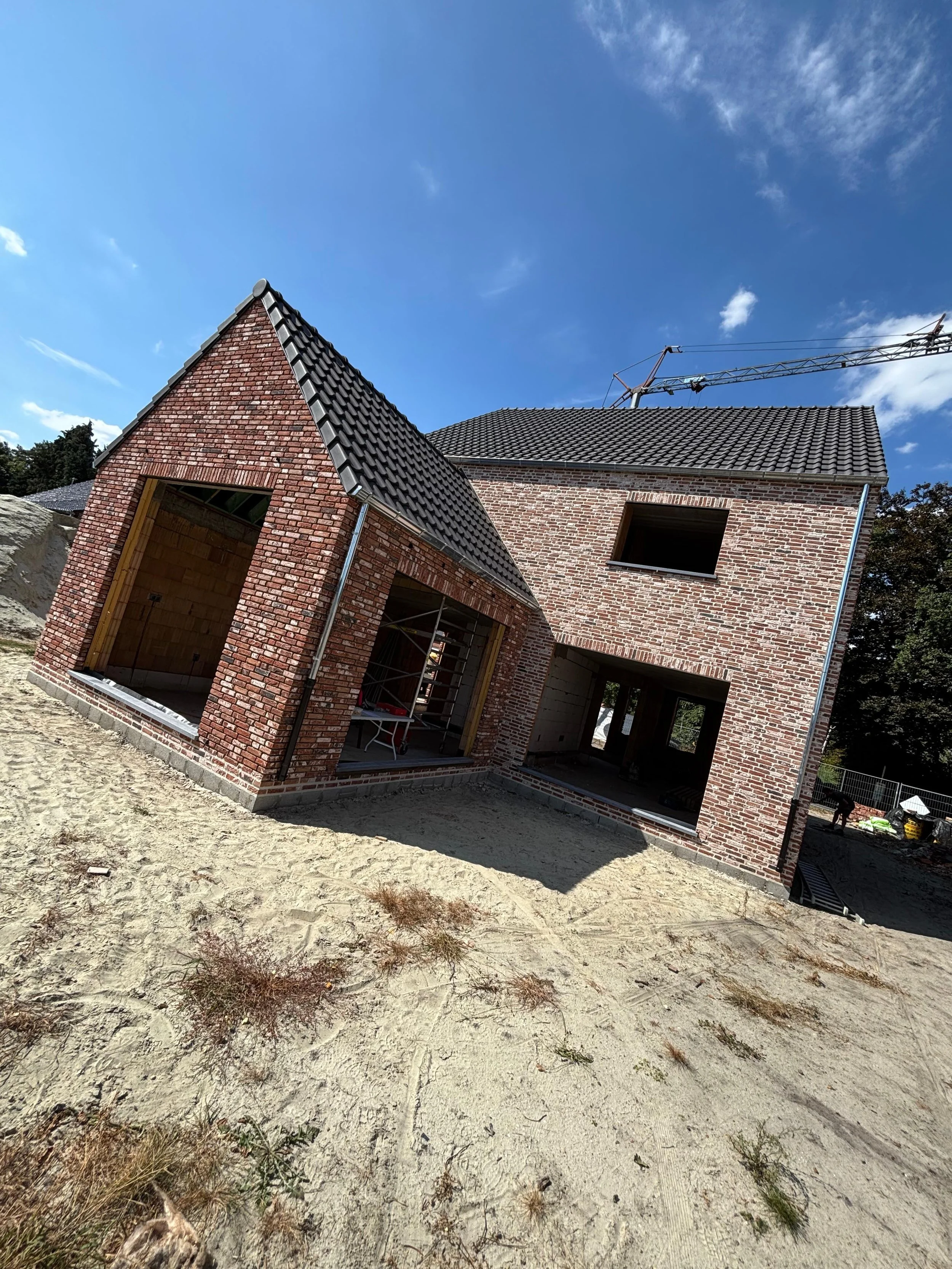 Een huis onder constructie met bakstenen muren en een hellend dak met dakpannen, op een bouwterrein met zand en planten.