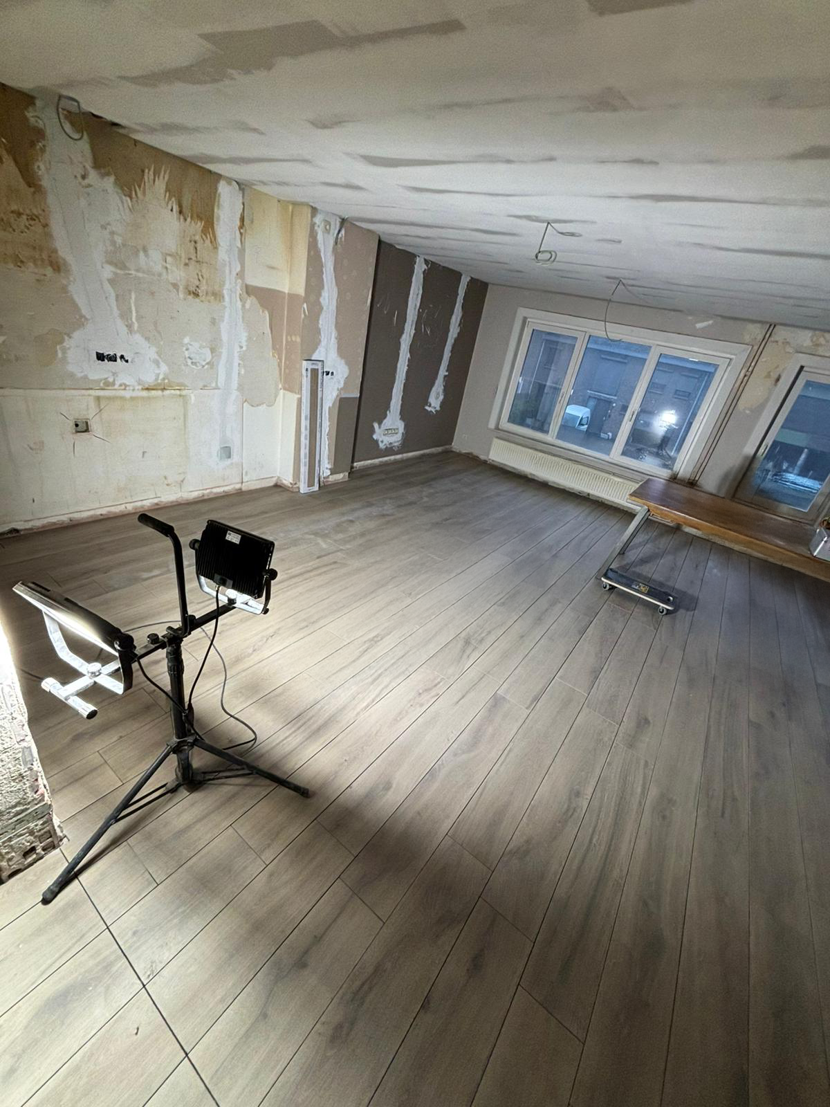 Een kamer in aanbouw met verouderde muren, nieuwe houten vloer, grote ramen, en bouwverlichting.