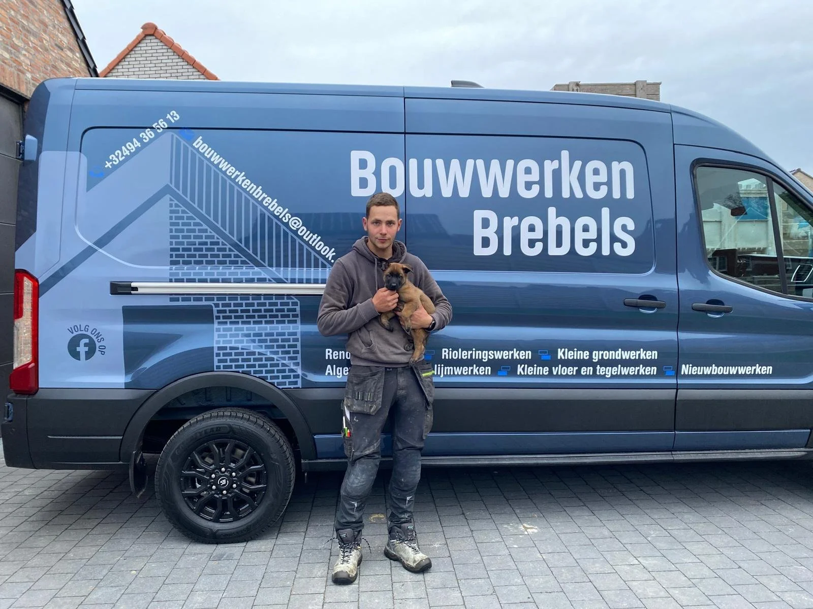 Man met een hond voor een blauwe vrachtwagen met bouwbedrijf branding, op een oprit.