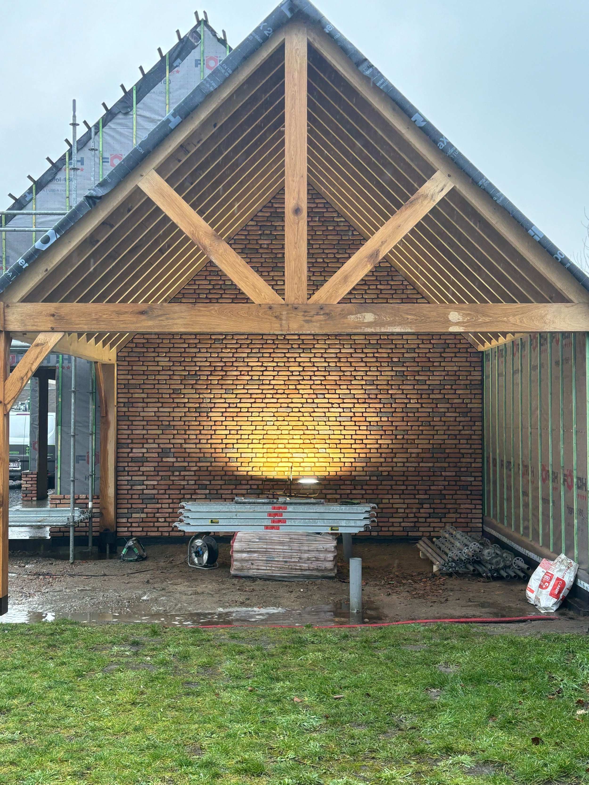 Een carport in aanbouw met een houten structuur en bakstenen muur, bouwmaterialen en gereedschap op de werf.