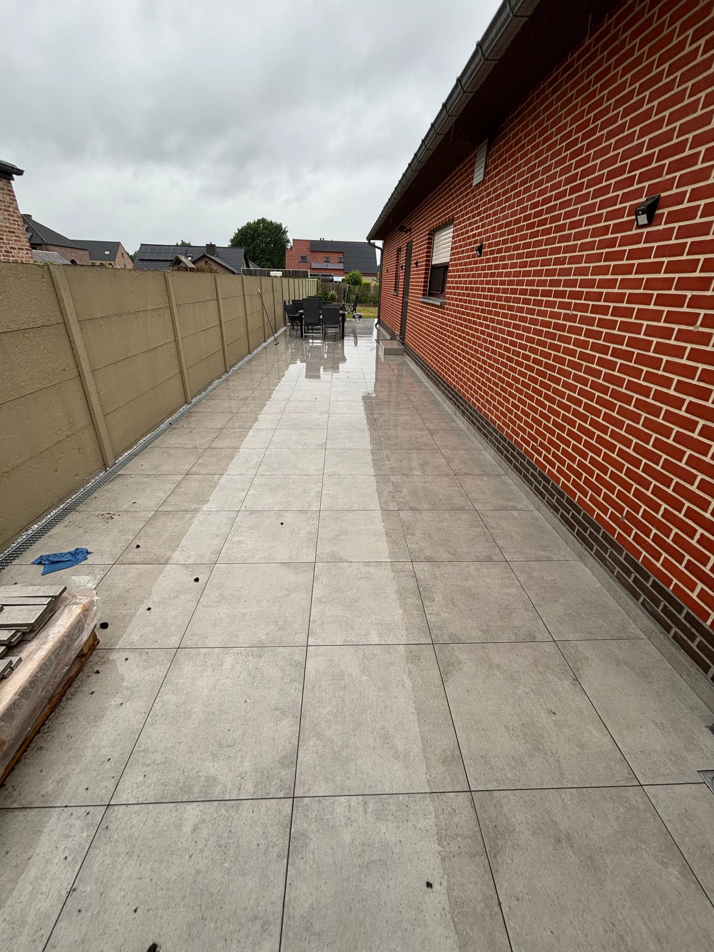 Een betegeld terras naast een rood bakstenen muur met buitenverlichting, met een tuintafel en stoelen op de achtergrond en een houten afzetting aan de linkerzijde.