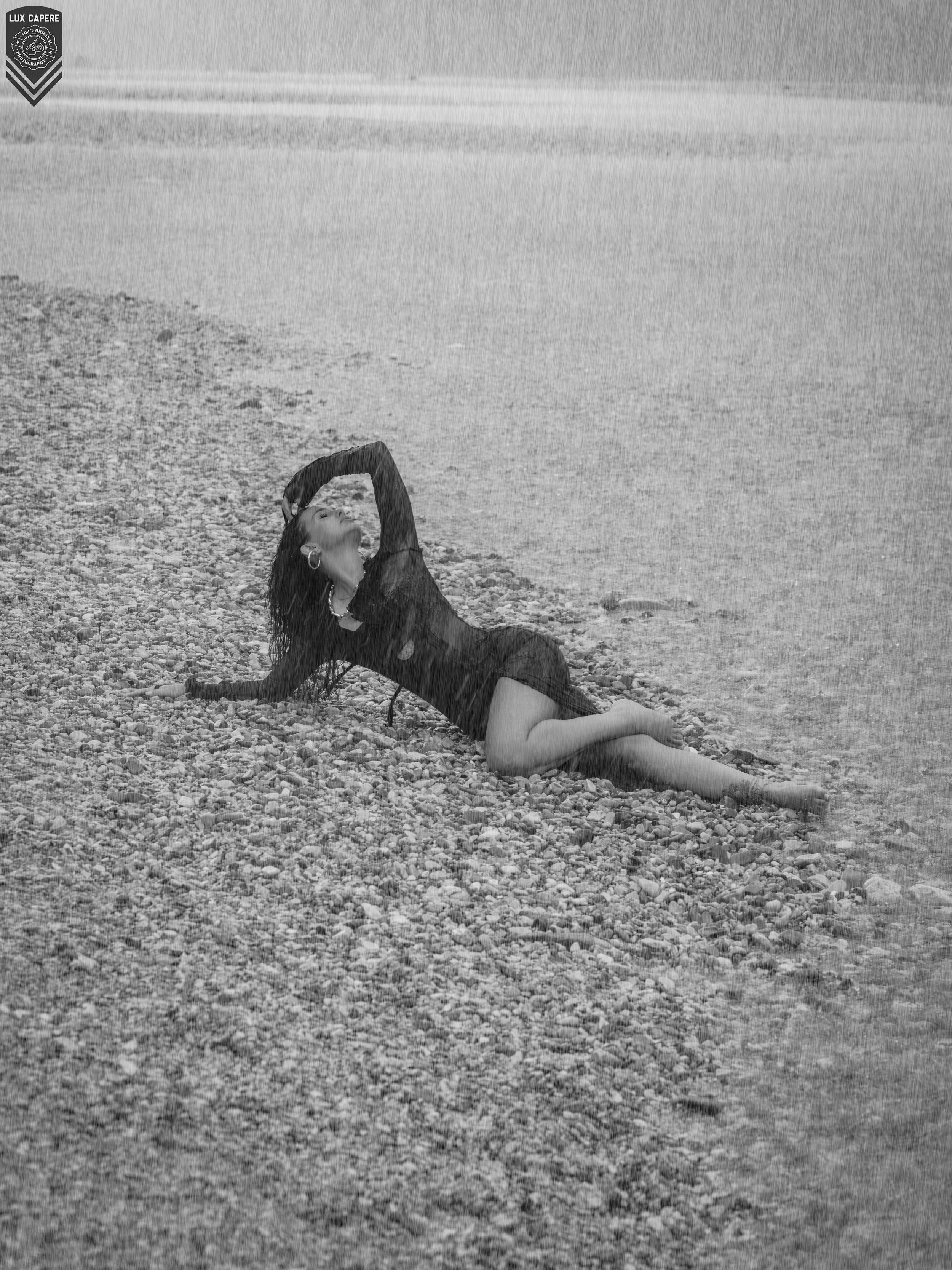 Frau liegt auf einem Kiesstrand, blickt nach oben, Wasser im Hintergrund, nasse Wassertropfen auf dem Bild. Portrait- und Cinematic-Fotografie von Lux Capere