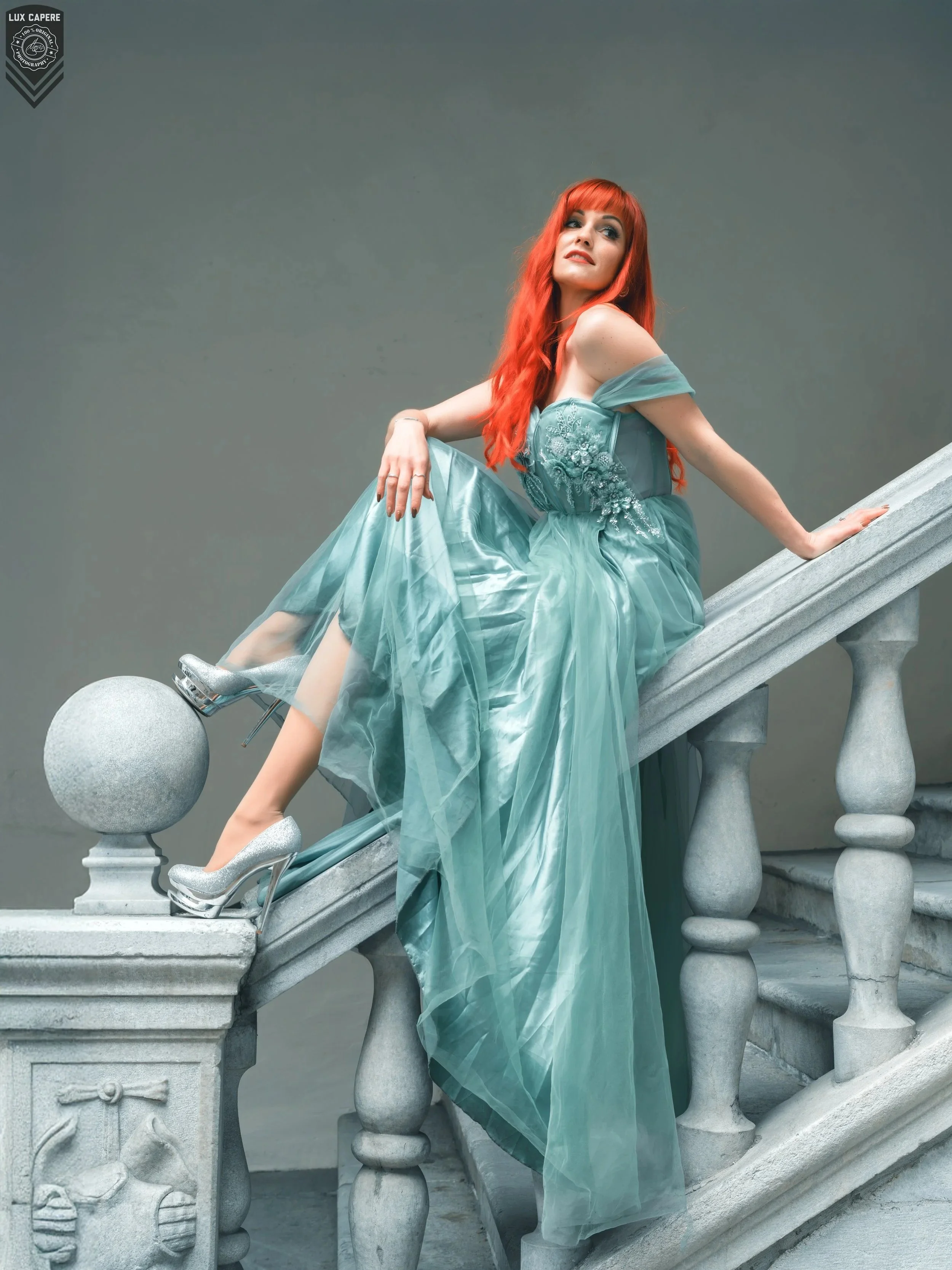 Eine Frau mit roten Haaren in einem eleganten, türkis-blauen Kleid sitzt auf einer Treppe aus Stein, vor einem grauen Hintergrund. Portrait- und Cinematic-Fotografie von Lux Capere