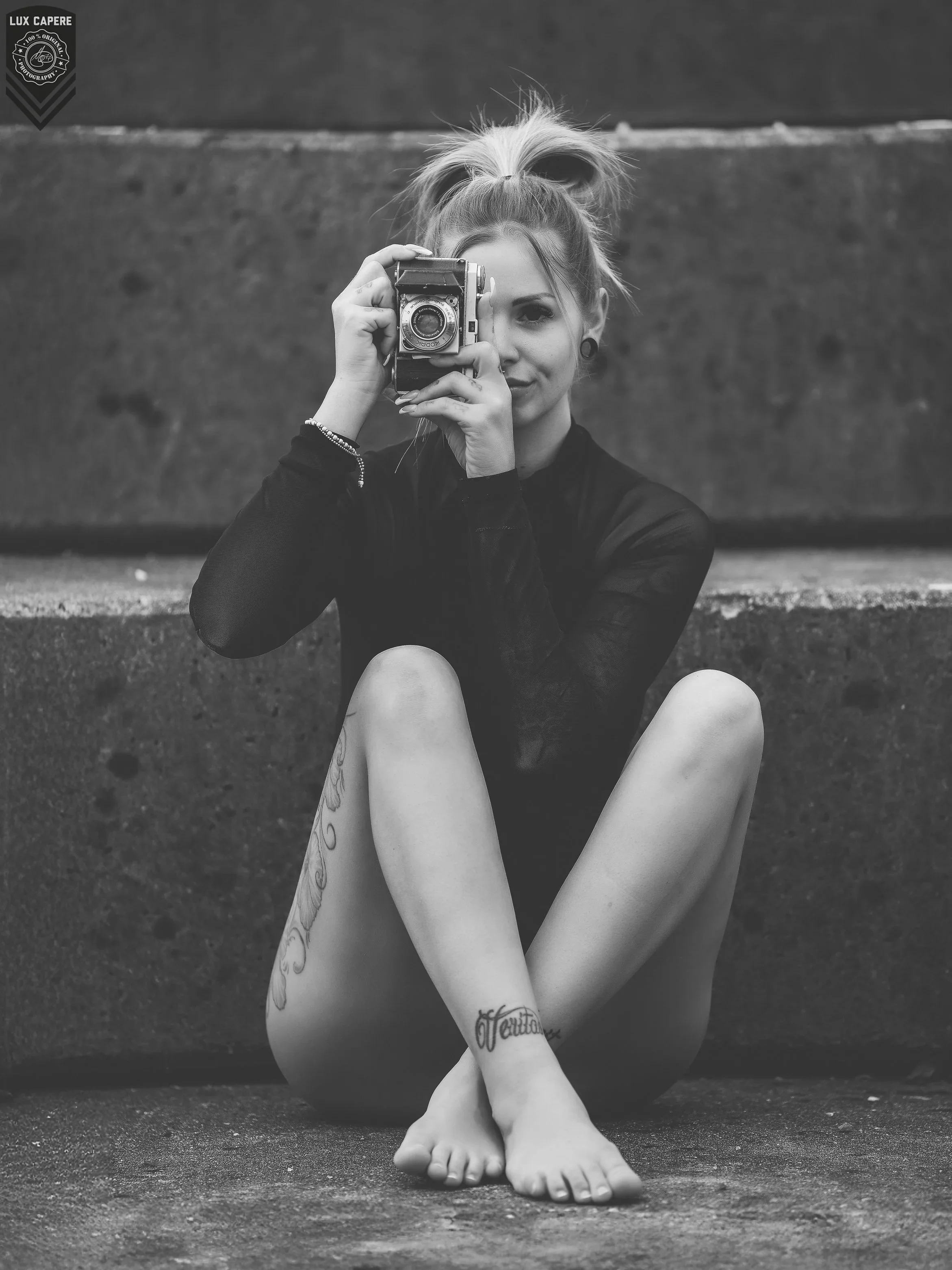 Schwarz-Weiß-Foto eines Mädchens, das auf dem Boden sitzt und eine Kamera vor das rechte Auge hält, mit Tattoos an den Beinen und Armbändern, vor einem dunklen Hintergrund. Portrait- und Cinematic-Fotografie von Lux Capere