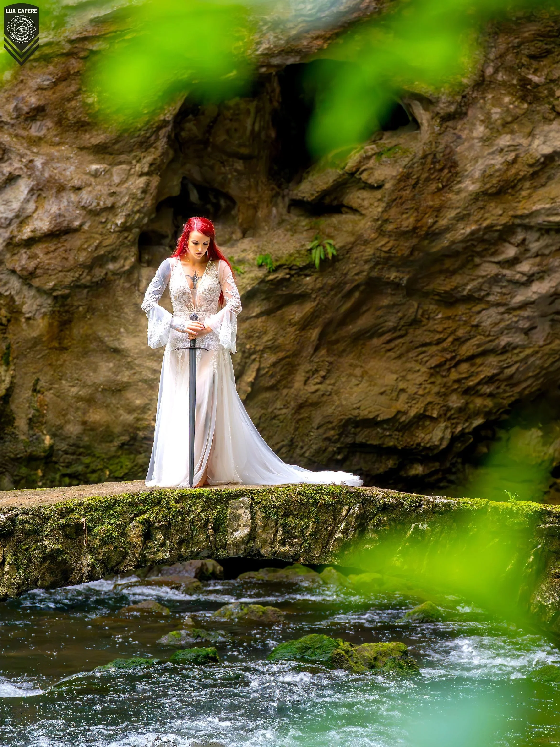 Eine Frau in einem weißen, eleganten Kleid mit roten Haaren steht am Rand eines Flusses, hält ein Schwert und blickt nach unten, umgeben von Felsen und grünem Wasser mit Blättern im Vordergrund. Portrait- und Cinematic-Fotografie von Lux Capere