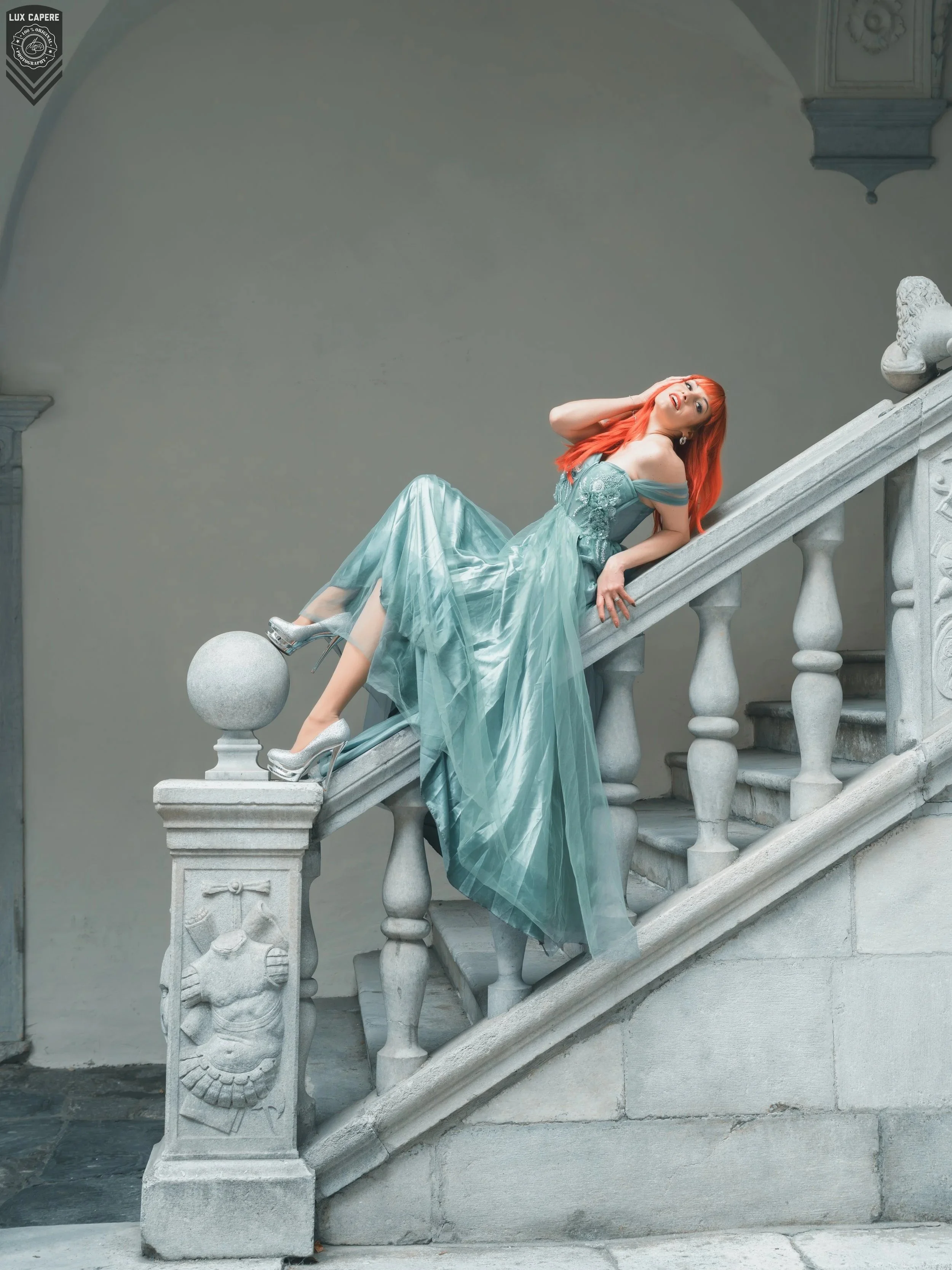 Frau mit roten Haaren in einem eleganten, hellblauen Kleid liegt auf einer steinernen Treppe und lehnt an der Brüstung. Portrait- und Cinematic-Fotografie von Lux Capere