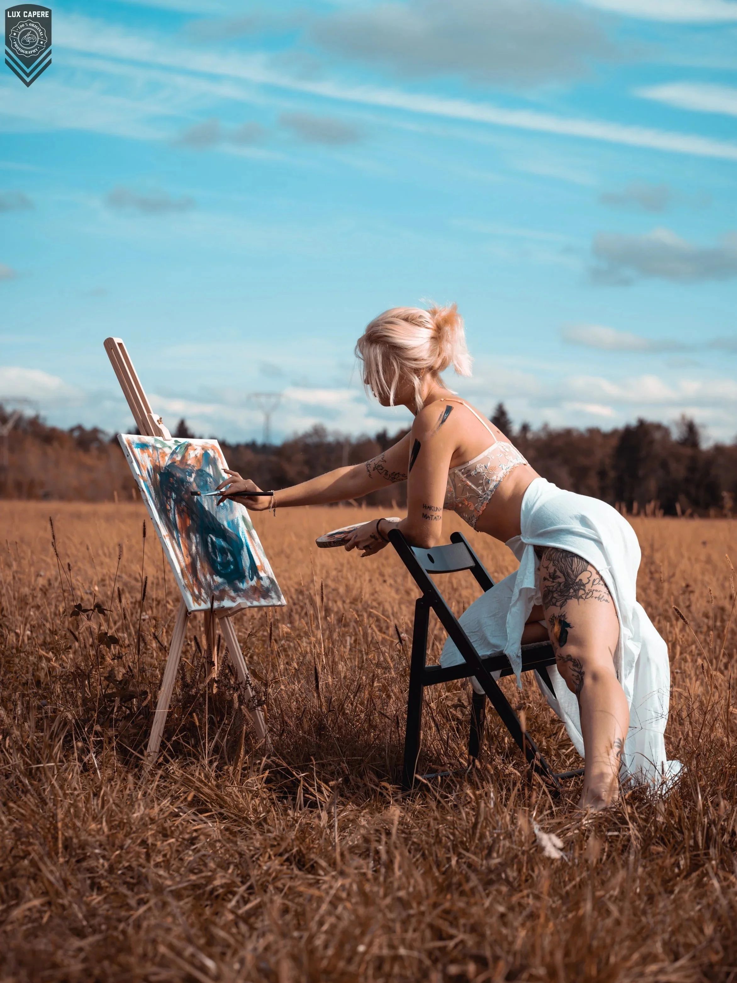 Junge Frau beim Malen im Freien auf einer Wiese , mit einem Easel und leuchtend blonden Haaren, trägt ein leichtes Tops und eine weiße Rock, viele Tattoos sichtbar. Portrait- und Cinematic-Fotografie von Lux Capere