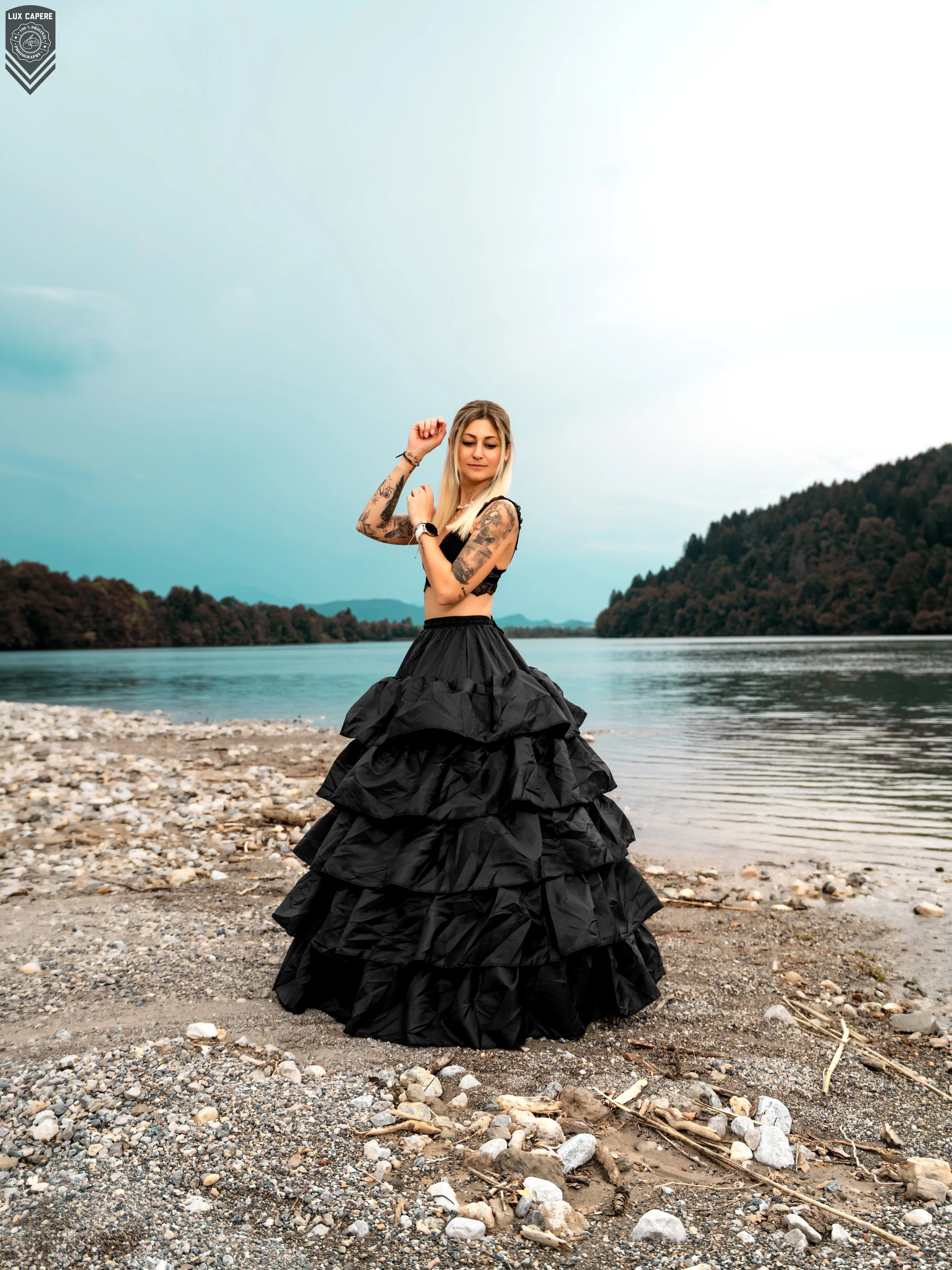 Junge Frau in einem schwarzen, ballkleidähnlichen Rock steht am Ufer eines Flusses, umgeben von Bäumen und Bergen im Hintergrund, mit einem bewölkten Himmel. Portrait- und Cinematic-Fotografie von Lux Capere