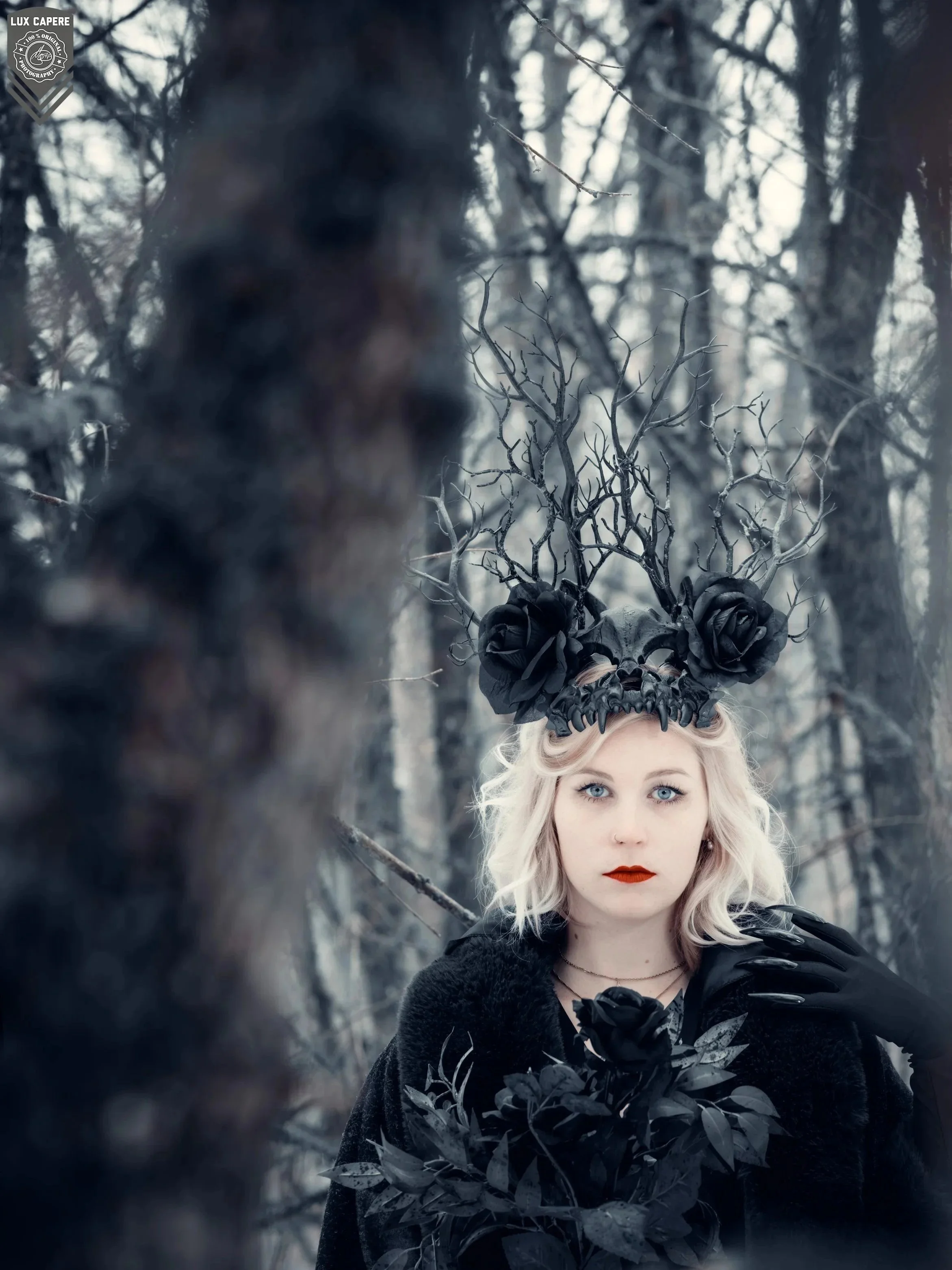Porträt einer jungen Frau mit blonden, welligen Haaren, rotem Lippenstift und schicker Kleidung, die in einem dunklen, winterlichen Wald steht, ergänzt durch ein kunstvolles, schwarzes Kopfstück aus Tierknochen und Ästen, das wie eine Krone aussieht.