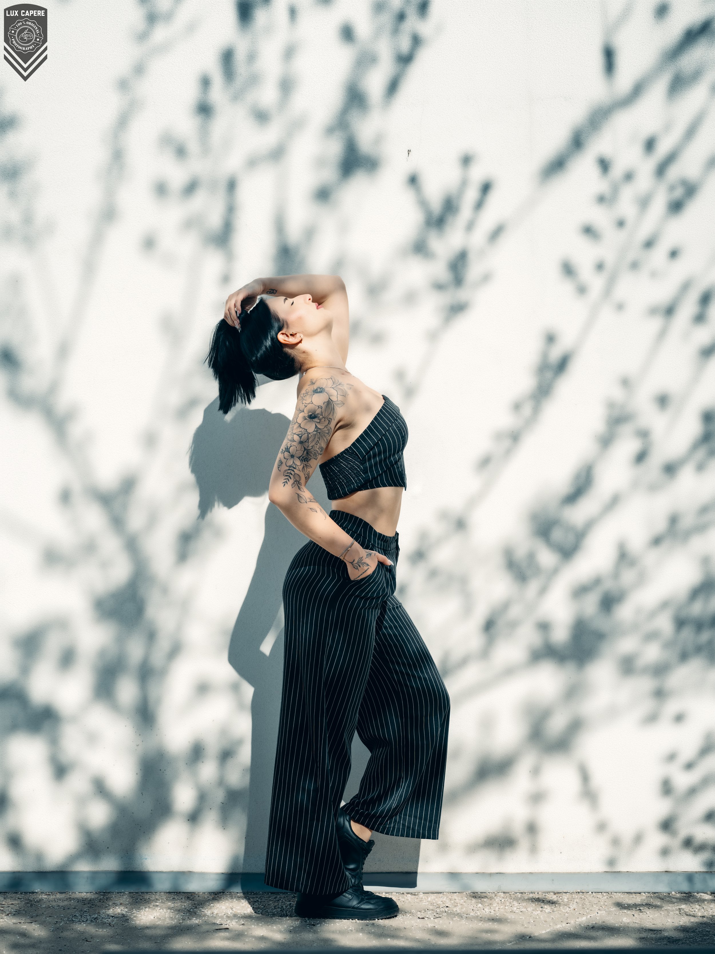 Junge Frau mit Tattoos trägt schwarze gestreifte Hose und Top, steht an einer weißen Wand mit Schatten von Ästen, pose mit Hand im Haar. Portrait- und Cinematic-Fotografie von Lux Capere