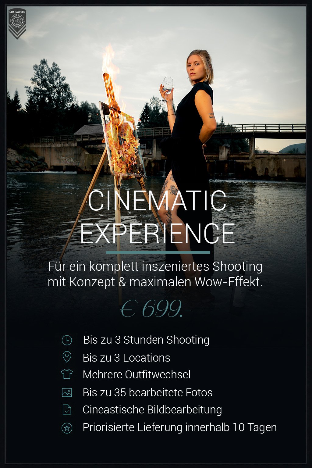 Junge Frau mit Tattoos in einem schwarzen Kleid, die neben einem brennenden Gemälde steht, das auf einem Holzständer befestigt ist, in einer Flusslandschaft mit Brücke im Hintergrund. Portrait- und Cinematic-Fotografie von Lux Capere.