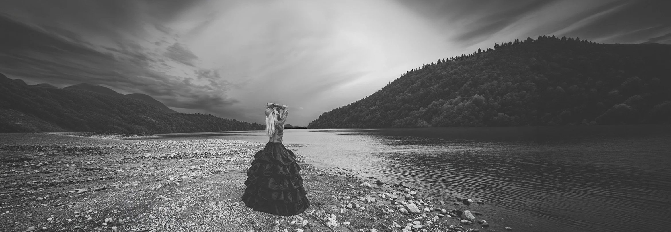 Eine Frau in einem langen schwarzen Rock steht an einem Flussufer vor einer Bergkulisse, die Himmel ist bewölkt, Schwarz-Weiß-Foto. Portrait- und Cinematic-Fotografie von Lux Capere