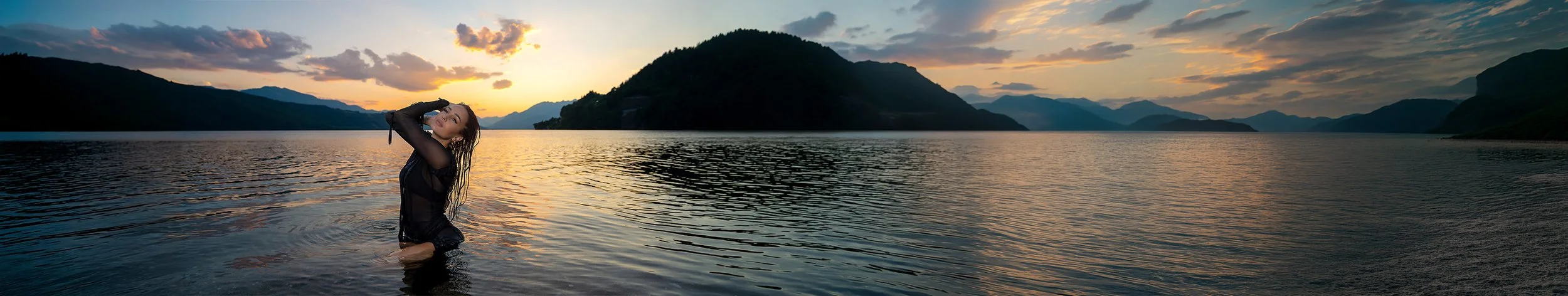 Eine Frau steht bis zu den Knien im Wasser eines Sees bei Sonnenuntergang, umgeben von Bergen und einem bewölkten Himmel. Portrait- und Cinematic-Fotografie von Lux Capere