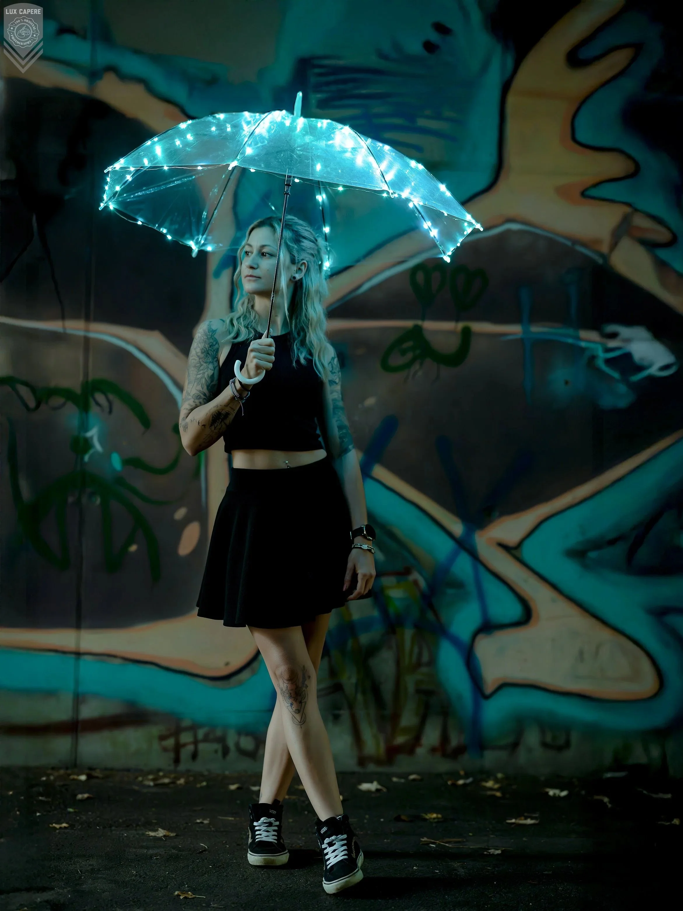 Eine junge Frau mit Tattoos und schwarzen Kleid, stehend vor einer bunten Graffiti-Wand, hält einen transparenten Regenschirm mit blinkenden Lichter. Sie trägt Turnschuhe und sieht nach links. Portrait- und Cinematic-Fotografie von Lux Capere