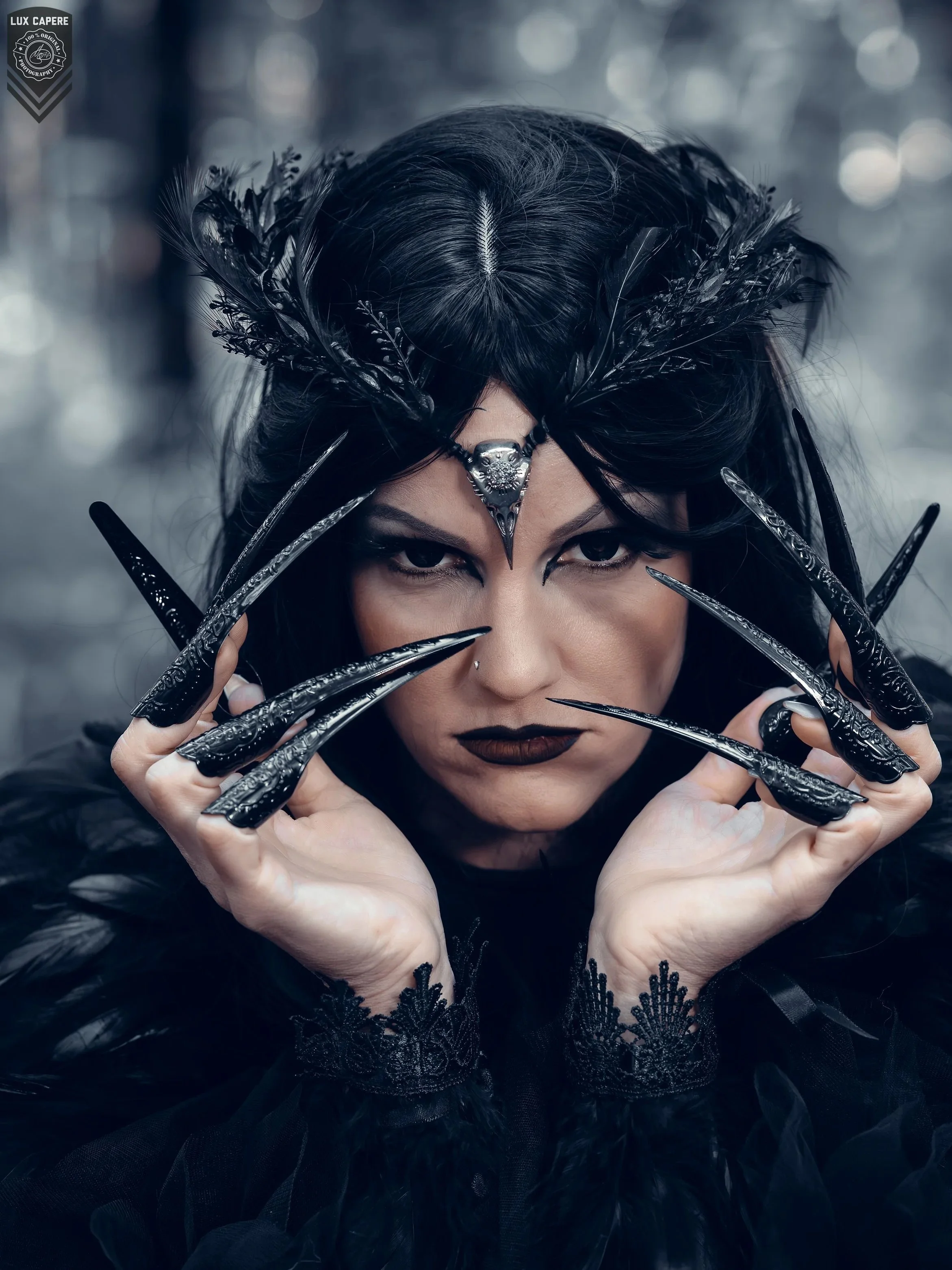 Nahaufnahme eines Gesichts einer Frau, die schwarz gekleidete, elegante Kleidung und Accessoires trägt. Sie hat dunkle Lippen, einen Piercing an der Unterlippe und trägt einen auffälligen schwarzen Kopfschmuck mit Federn und einem silbernen Schmuckstück auf der Stirn. Ihre Hände, die mit schwarzen, kunstvoll gestalteten Fingernägeln bedeckt sind, sind nahe an ihrem Gesicht positioniert.