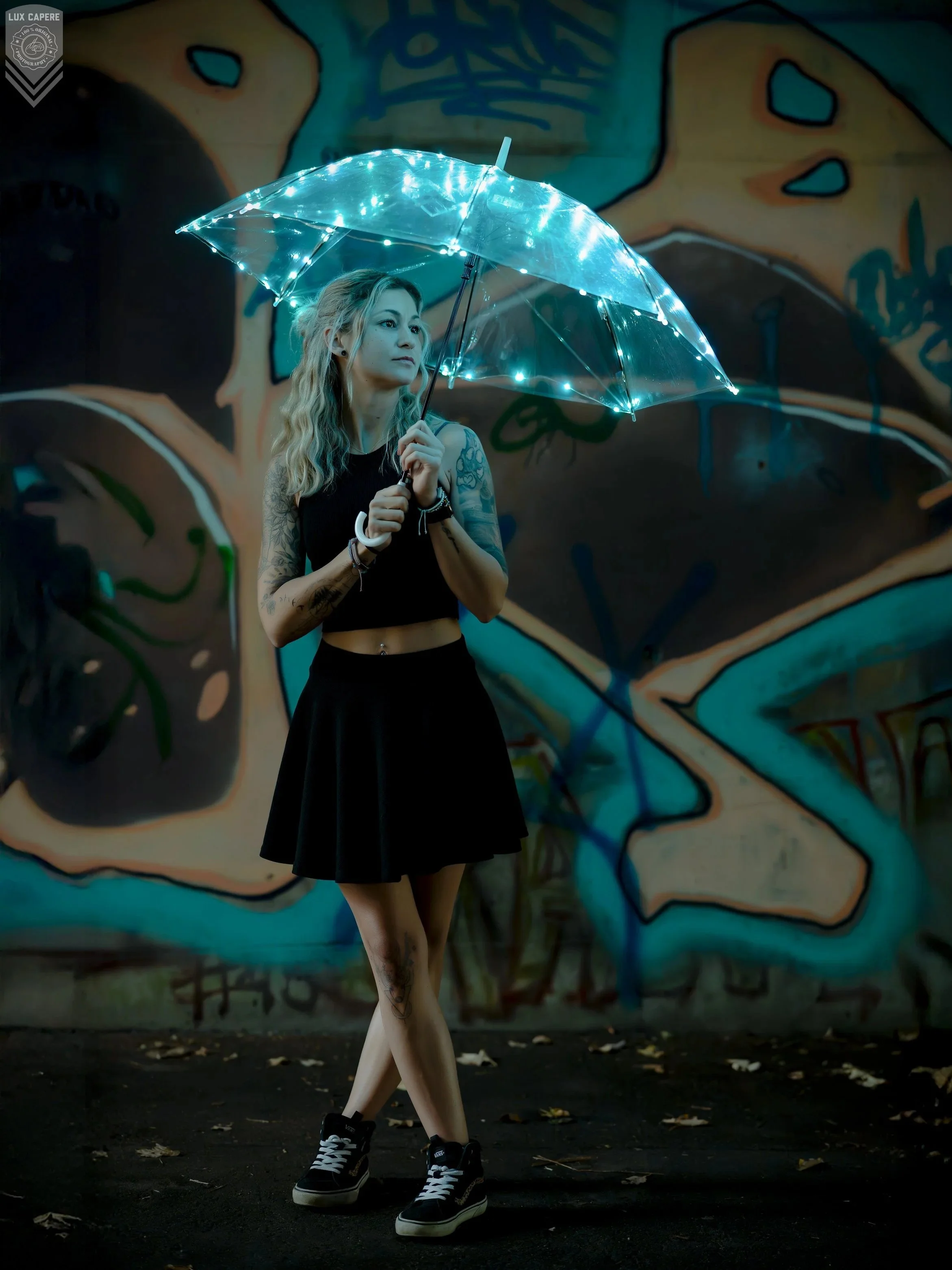 Junge Frau mit Tattoos trägt einen schwarzen Rock, schwarze Schuhe und hält einen leuchtenden, transparenten Regenschirm in der Hand. Hinter ihr befindet sich eine Graffiti-Wand. Portrait- und Cinematic-Fotografie von Lux Capere