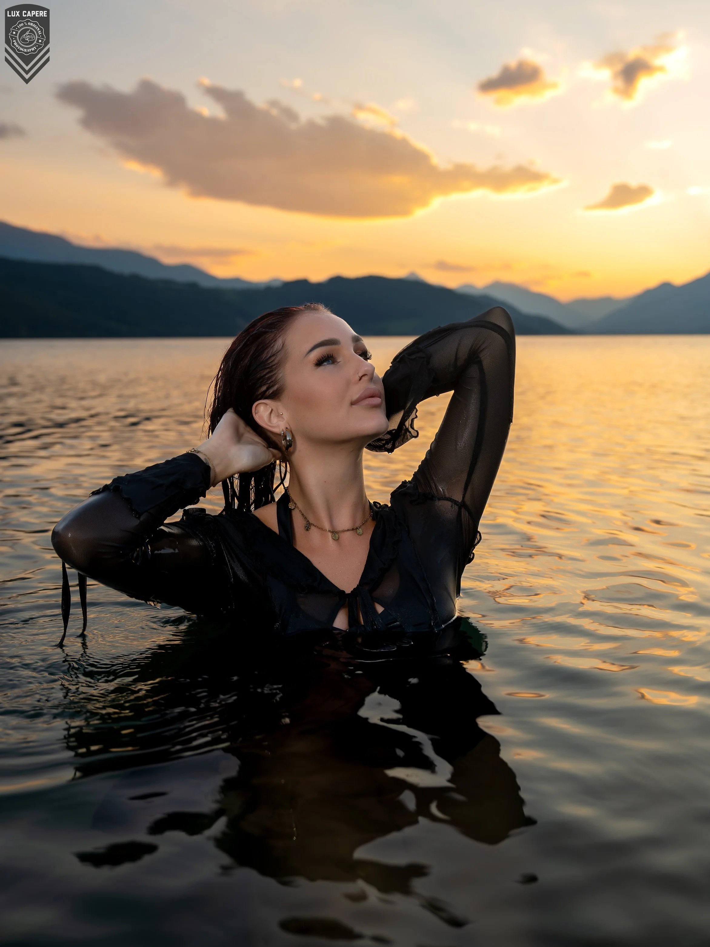 Eine junge Frau steht im Wasser eines Sees bei Sonnenuntergang, trägt schwarzes, durchsichtigen Top und schaut verträumt in den Himmel, umgeben von Bergen und einem Himmel mit Wolken, die im warmen Licht leuchten.