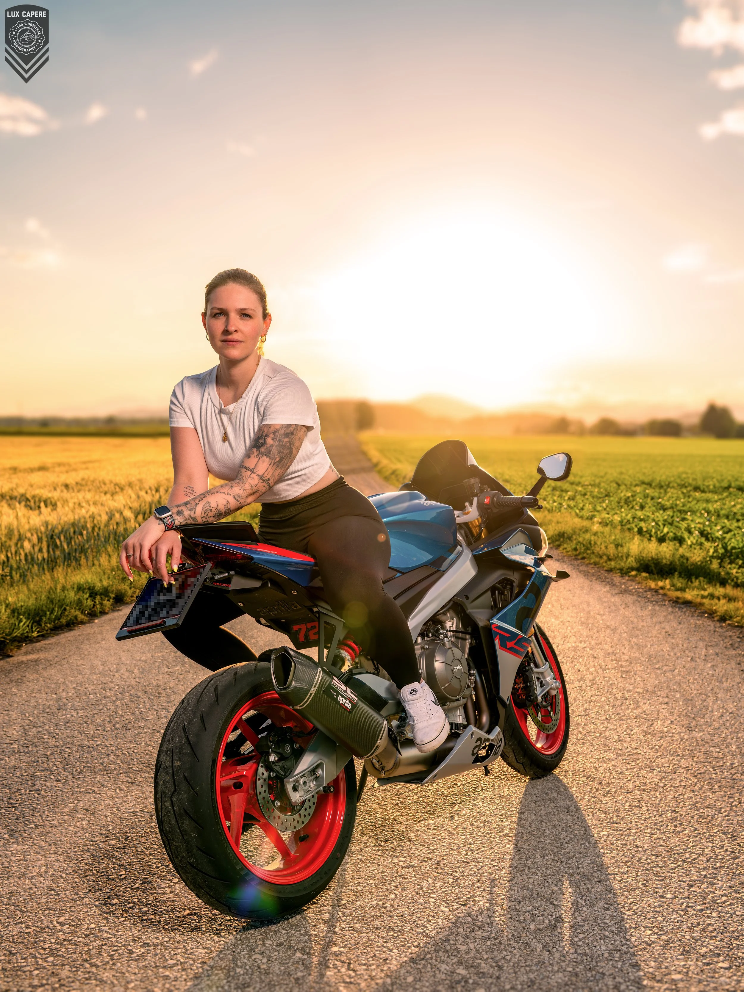 Junge Frau mit Tattoos sitzt auf einer sportlichen schwarzen und blauen Motorrad in einer ländlichen Landschaft bei Sonnenuntergang, während sie in die Kamera schaut. Portrait- und Cinematic-Fotografie von Lux Capere