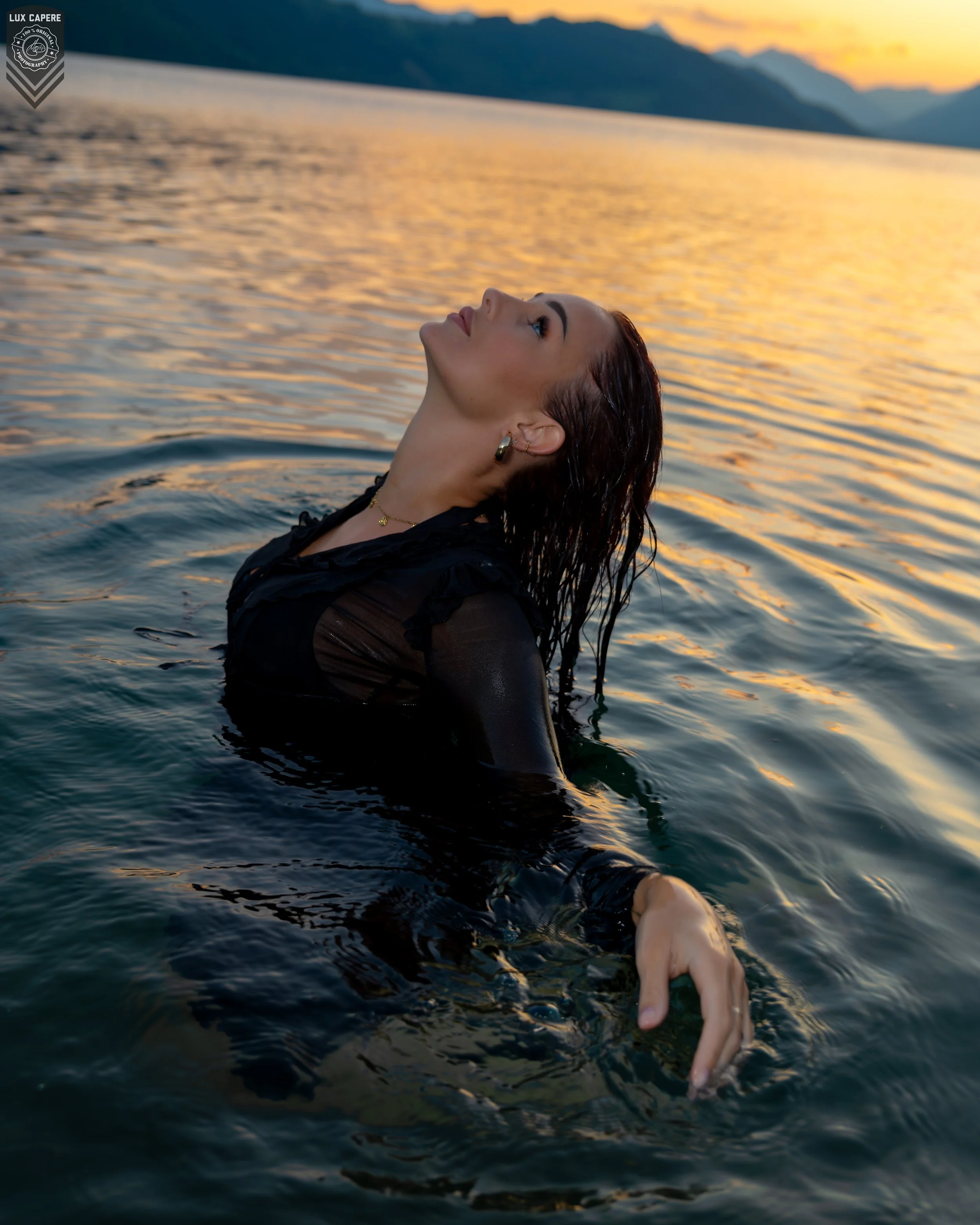 Frau im Wasser bei Sonnenuntergang, mit Wasser bis zum Hals, Blick nach oben, dunkle Kleidung, Berge im Hintergrund, Portrait- und Cinematic-Fotografie von Lux Capere
