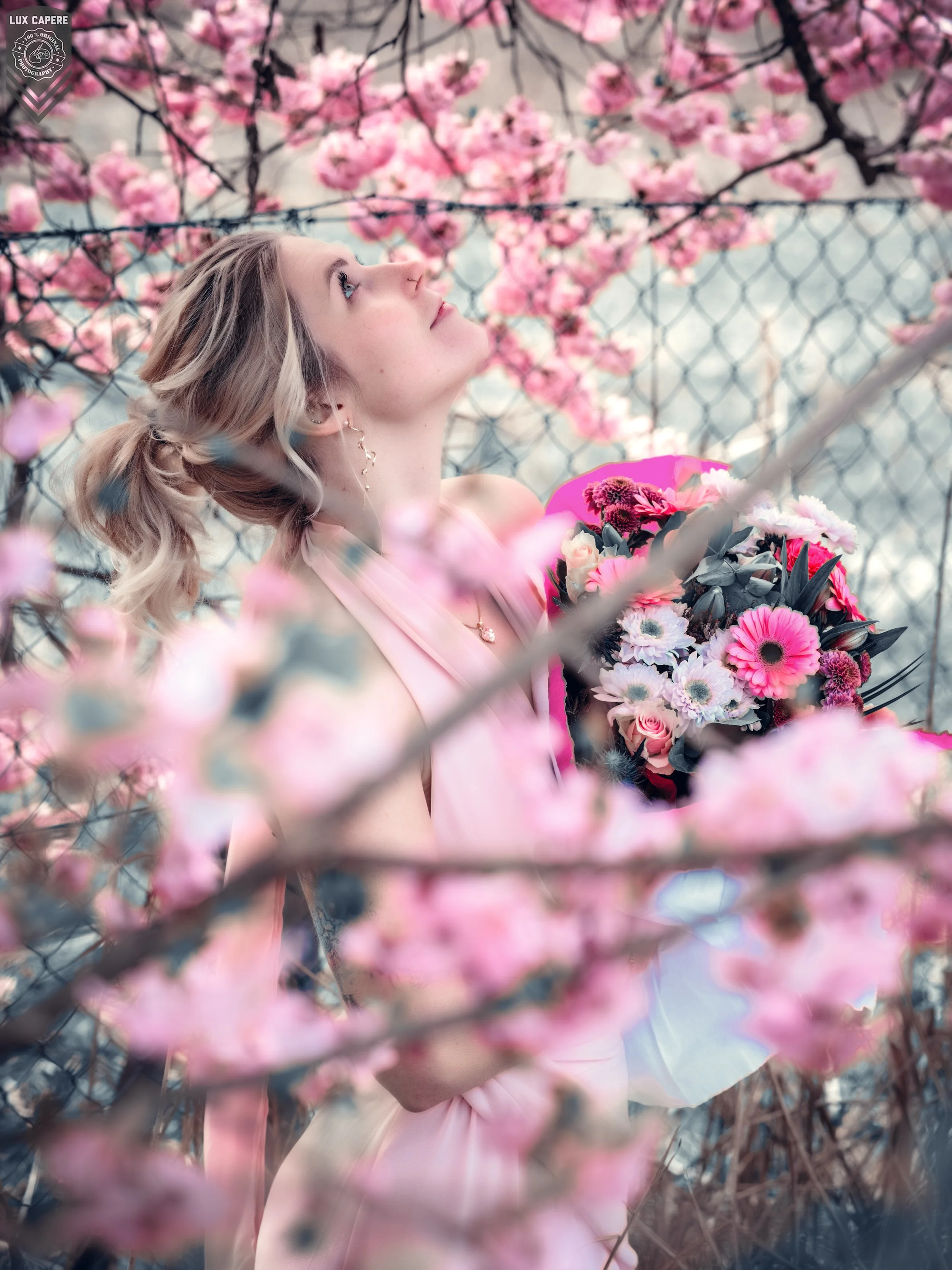 Junge Frau mit blondem, lockigem Haar hält einen Blumenstrauß aus rosa, weißen und roten Blumen mit pinken Blüten. Sie schaut nach oben, umgeben von blühenden Zweigen. Portrait- und Cinematic-Fotografie von Lux Capere