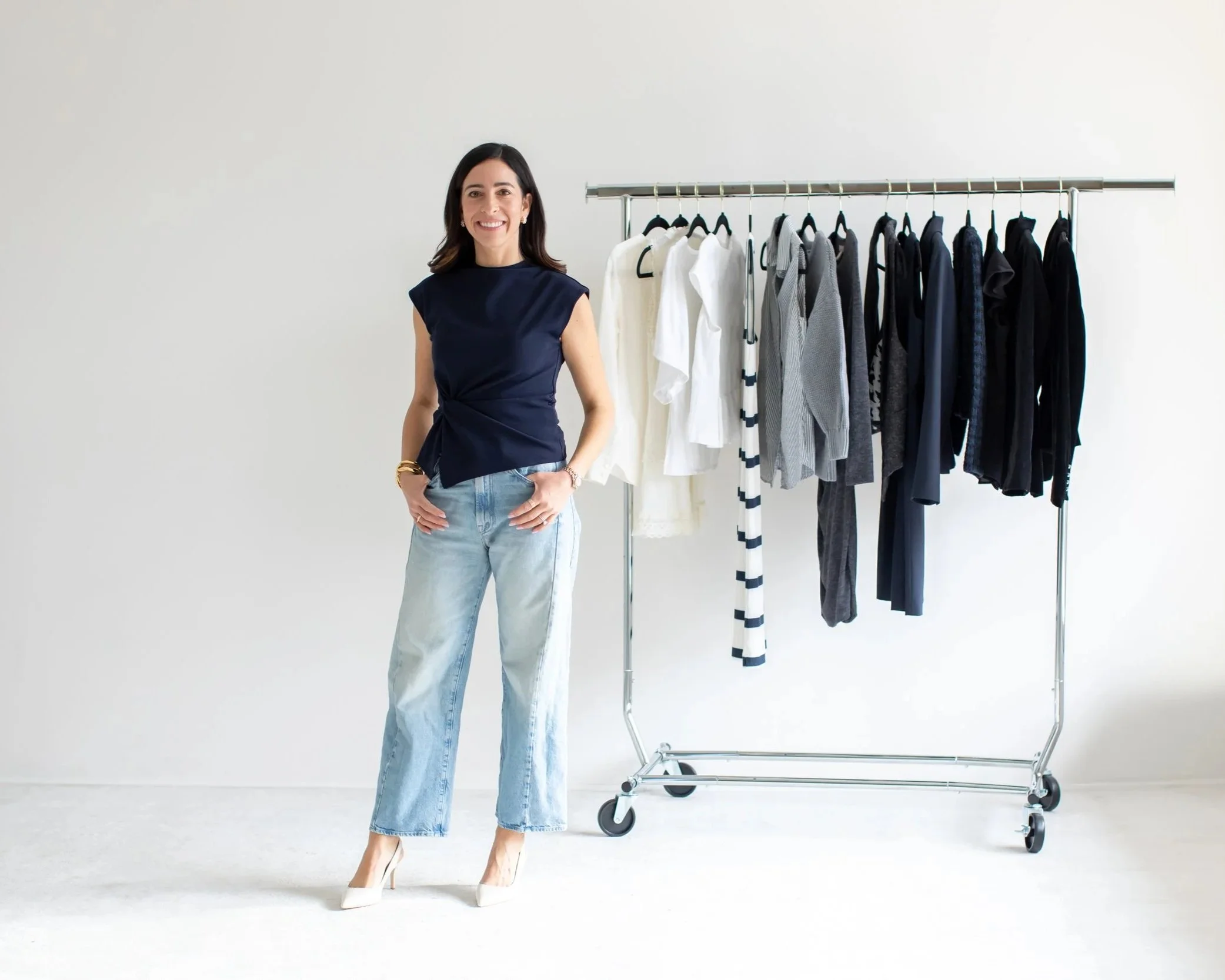 A woman standing next to a clothing rack with assorted white, gray, navy, and black garments in a bright room.