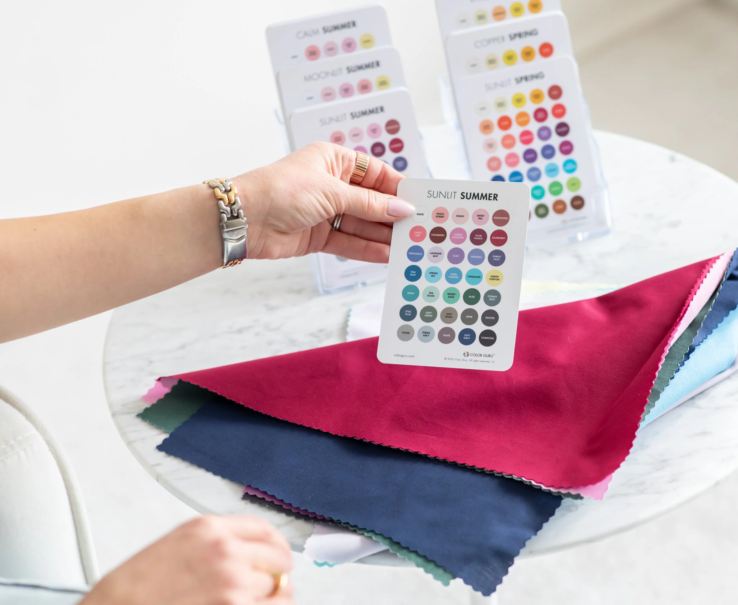 A person's hand holds a color sample card labeled "Sunlit Summer" with color names and shades, with fabric swatches in pink, navy blue, green, and other colors on a white marble table. In the background are multiple color sample cards displayed upright.