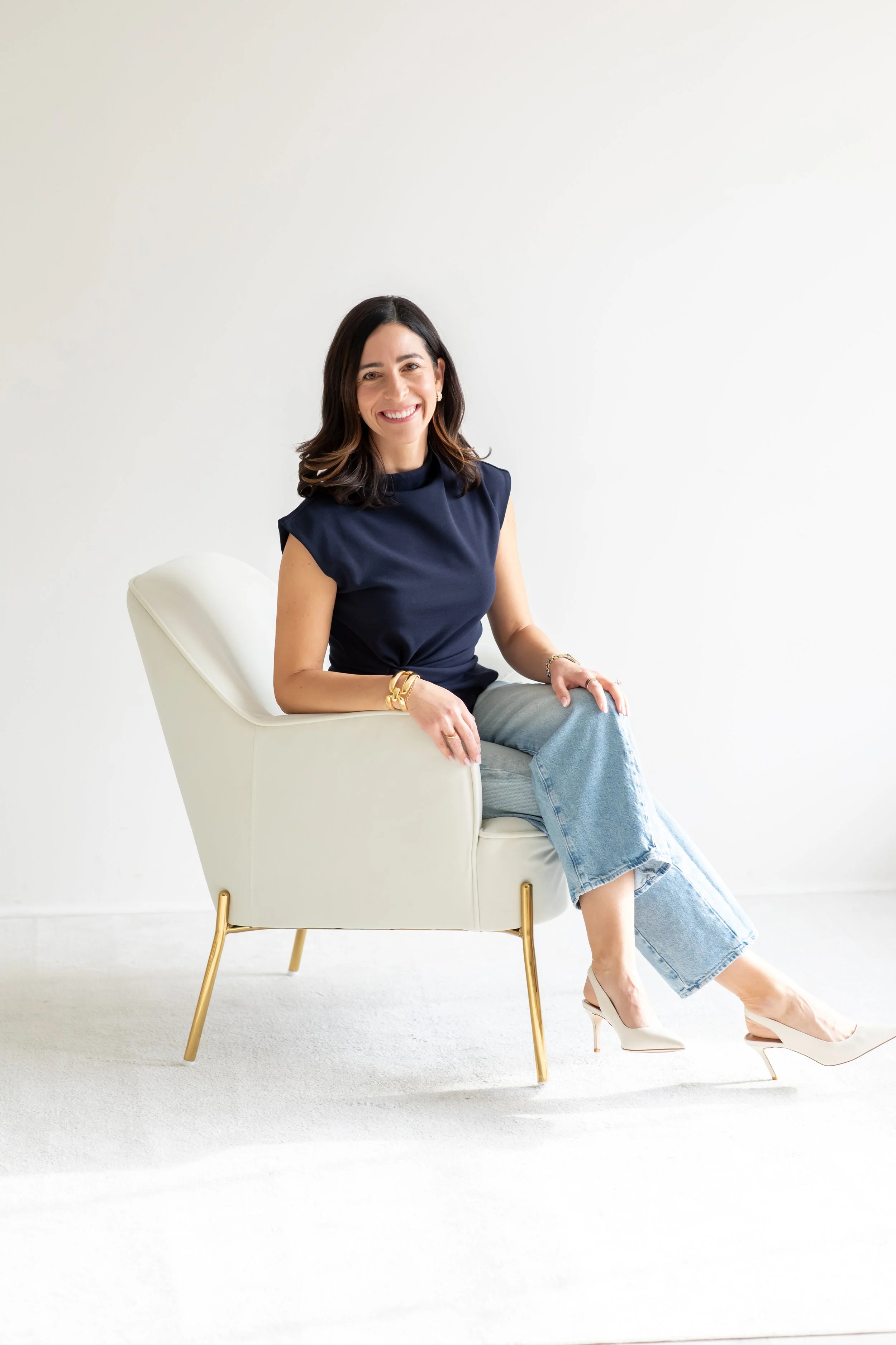 A woman sitting on a white chair with gold legs, wearing a navy sleeveless top, light blue jeans, and beige high heels, smiling.