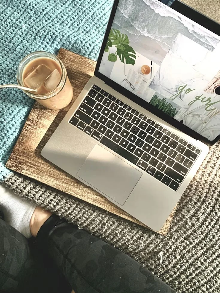 A laptop on a wooden tray with a glass of iced coffee or iced tea next to it, surrounded by a textured blue and gray rug, with a person's leg visible at the bottom.