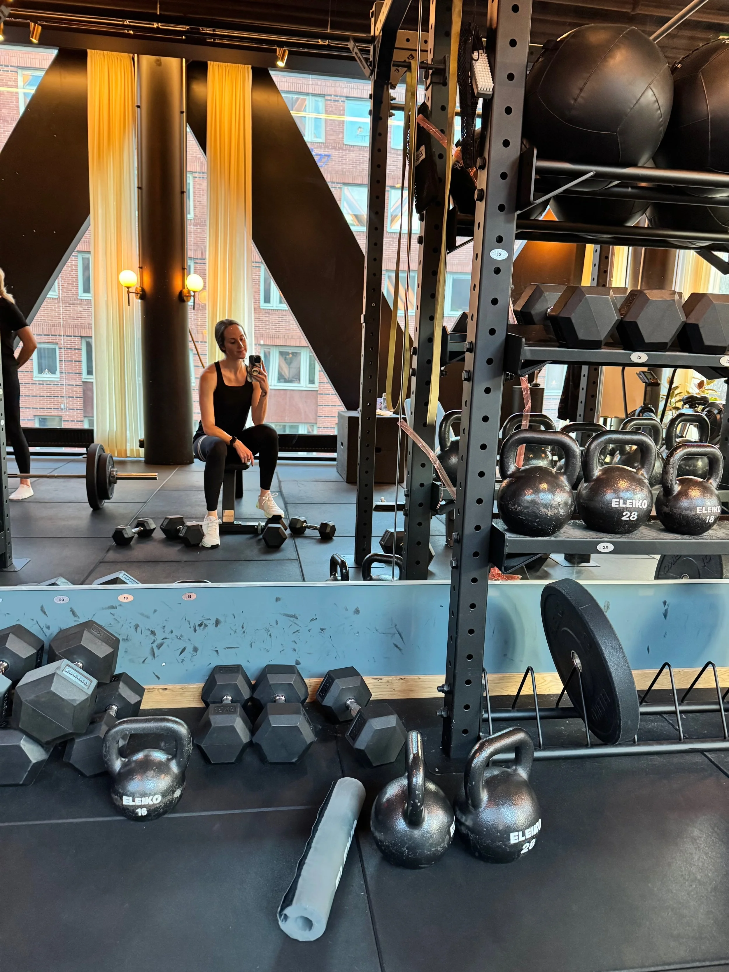 A gym with various weights, including dumbbells and kettlebells, with a woman sitting on a bench taking a selfie in the mirror. There are large windows with orange and yellow curtains and a reflection of a city building outside.