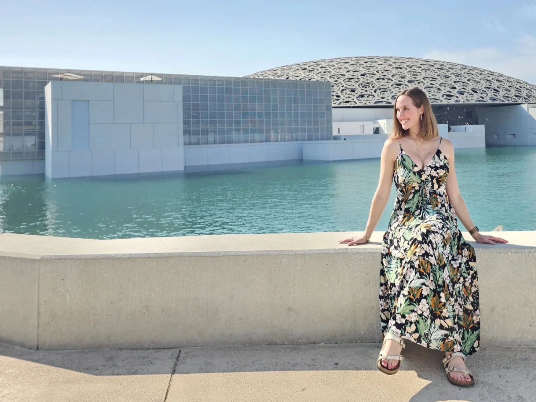 A woman in a floral dress sitting on a concrete ledge by a body of water with modern architecture in the background.