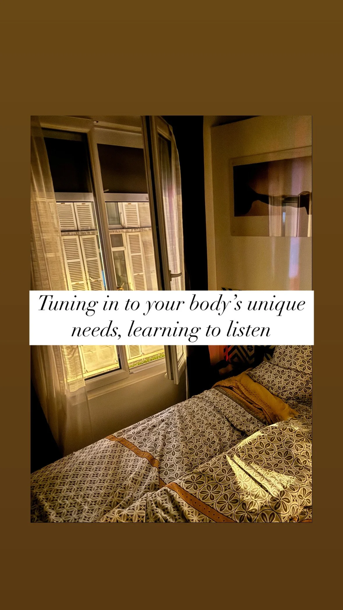 Sunlight coming through open window with curtains, view of city building with shutters, bed with patterned linens in a warmly lit room.