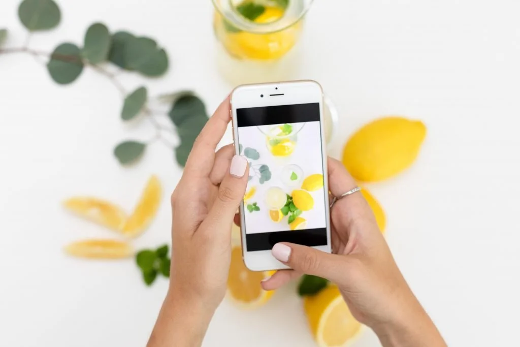 Person taking a photo of lemons, lemon slices, and eucalyptus leaves on a white surface with a smartphone.