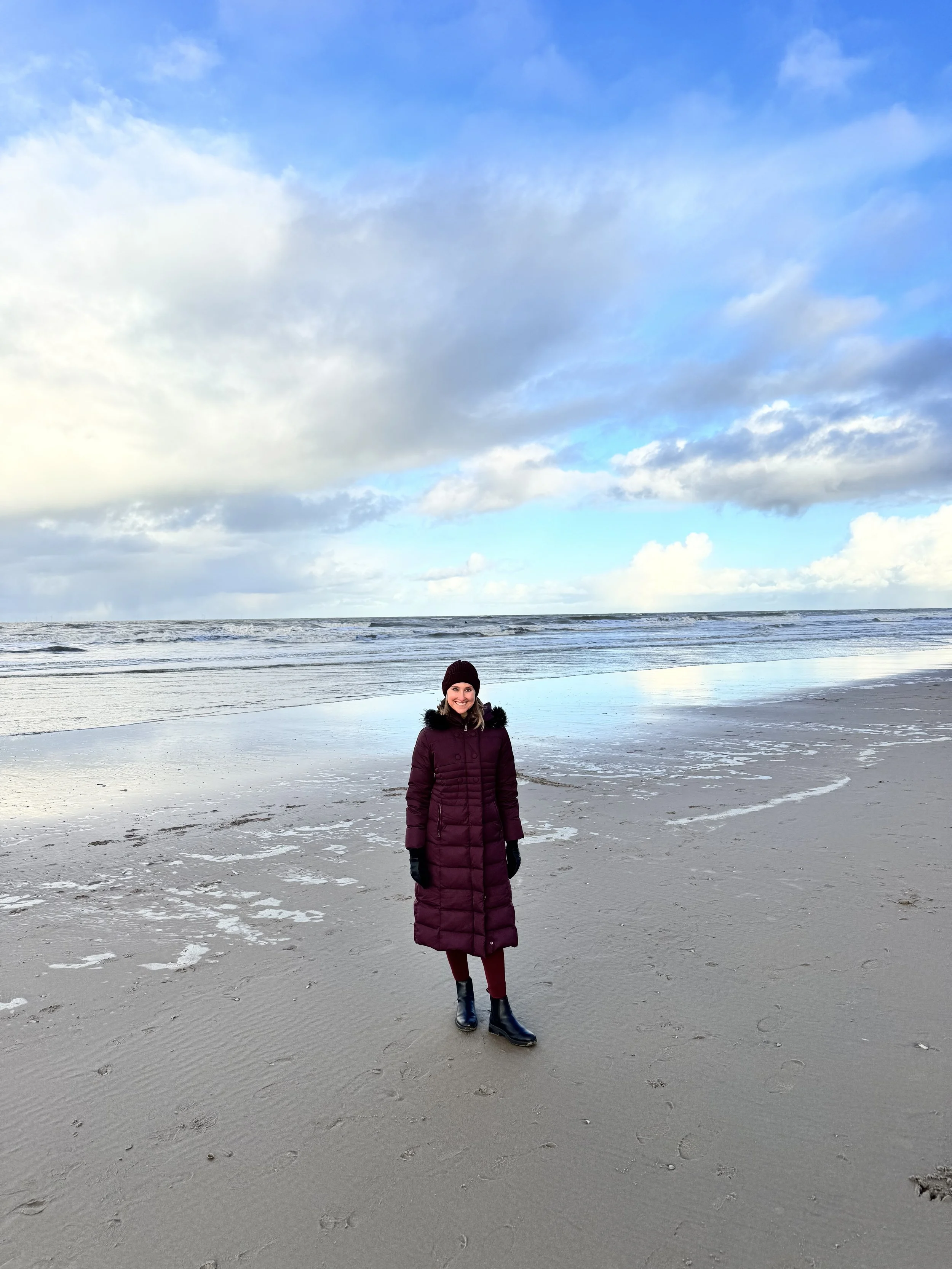 Woman in a burgundy winter coat, black gloves, and black boots standing on a sandy beach by the ocean with a cloudy blue sky overhead.