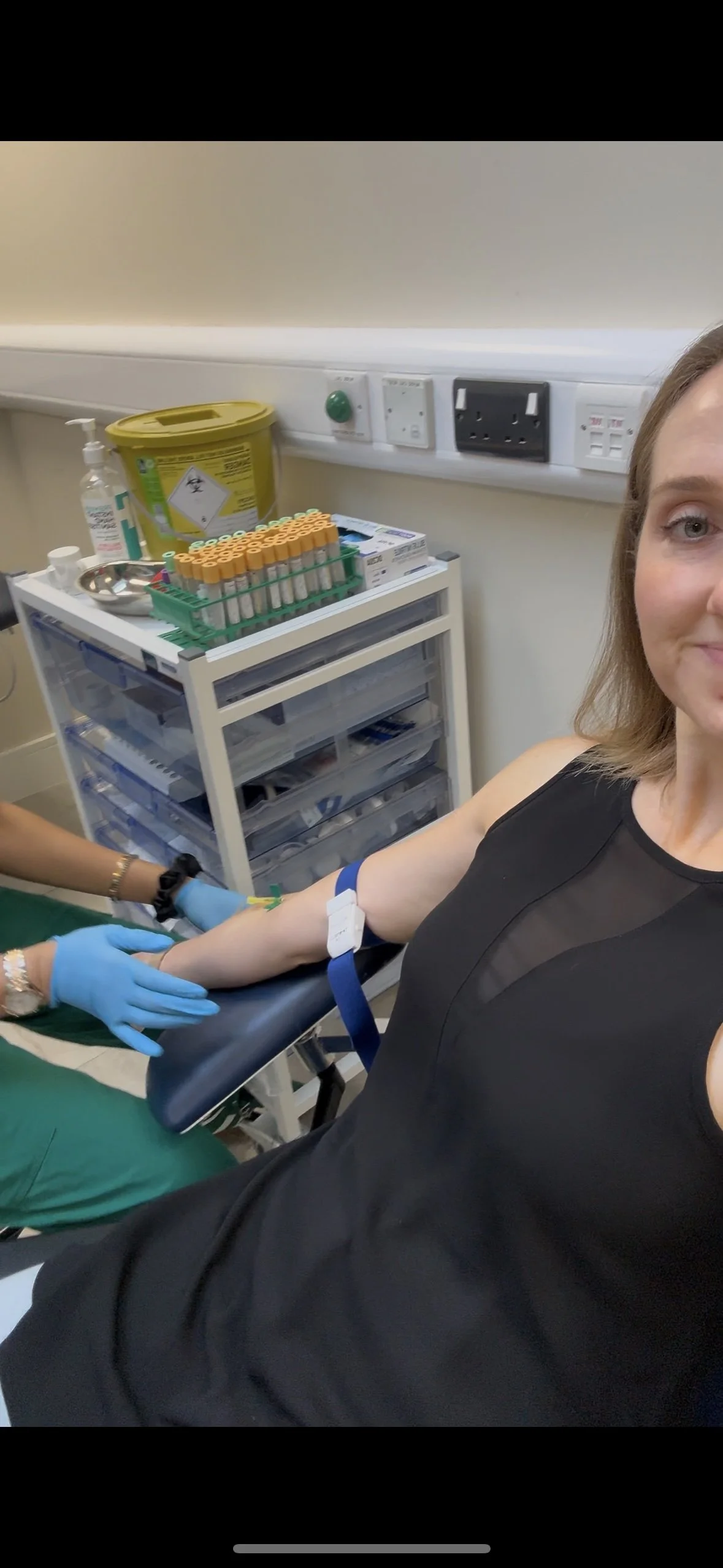 A woman donating blood in a medical room, with a healthcare worker preparing blood samples on a cart nearby.