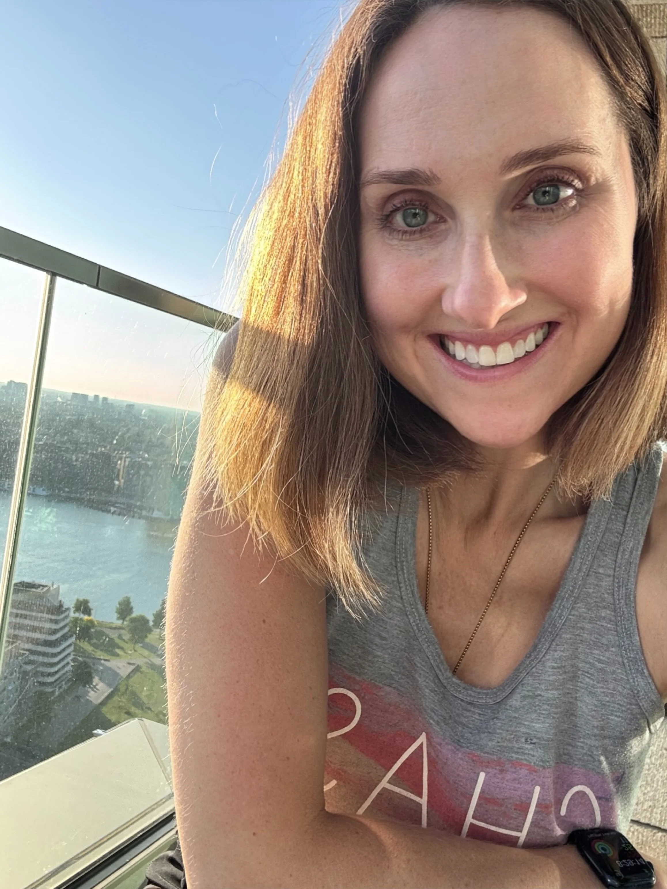 A young woman with shoulder-length brown hair and blue eyes smiling at the camera, standing on a balcony with a cityscape and water in the background during sunset.