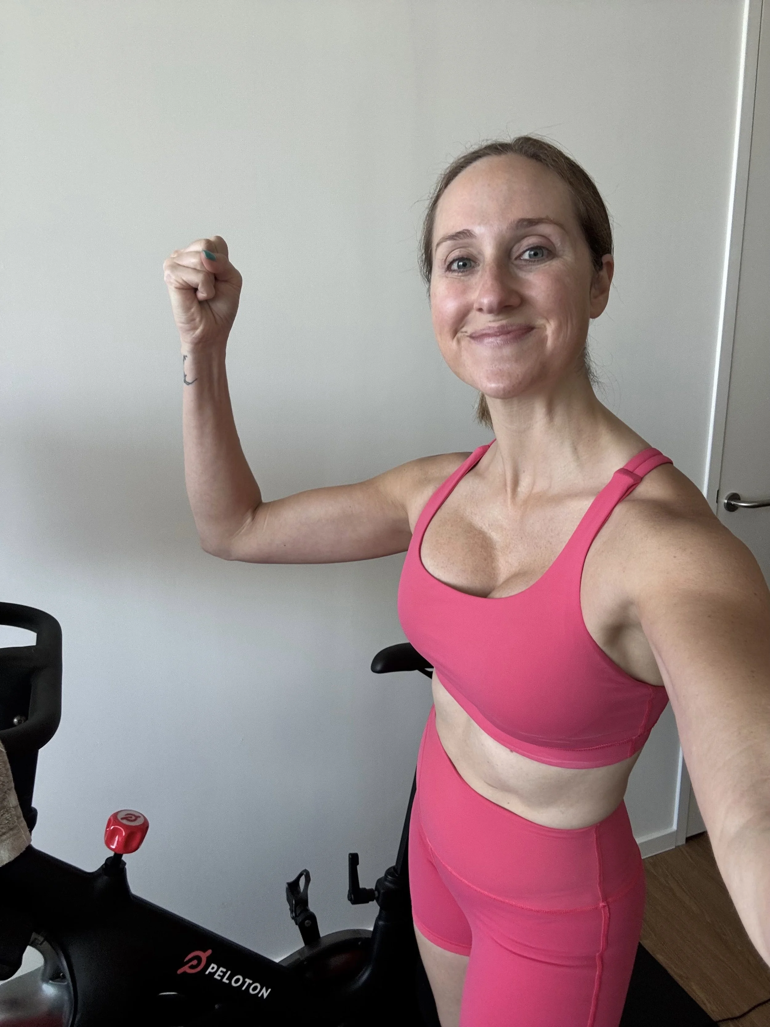 A woman in pink workout attire flexing her arm and smiling in a home gym with an exercise bike.