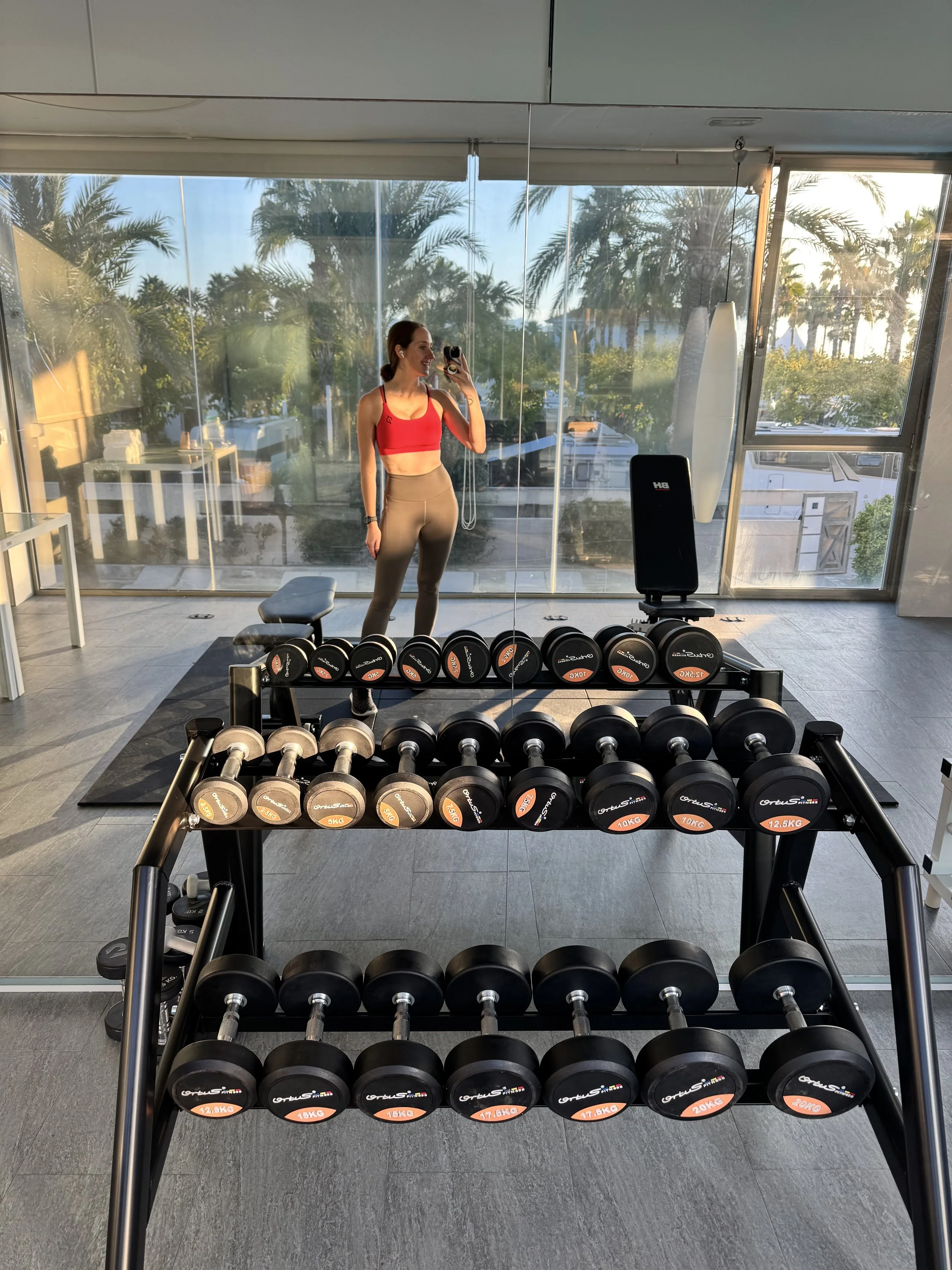 A woman taking a selfie in a gym, standing in front of a mirror with sunlight coming through large windows, with a rack of various sized dumbbells in the foreground.