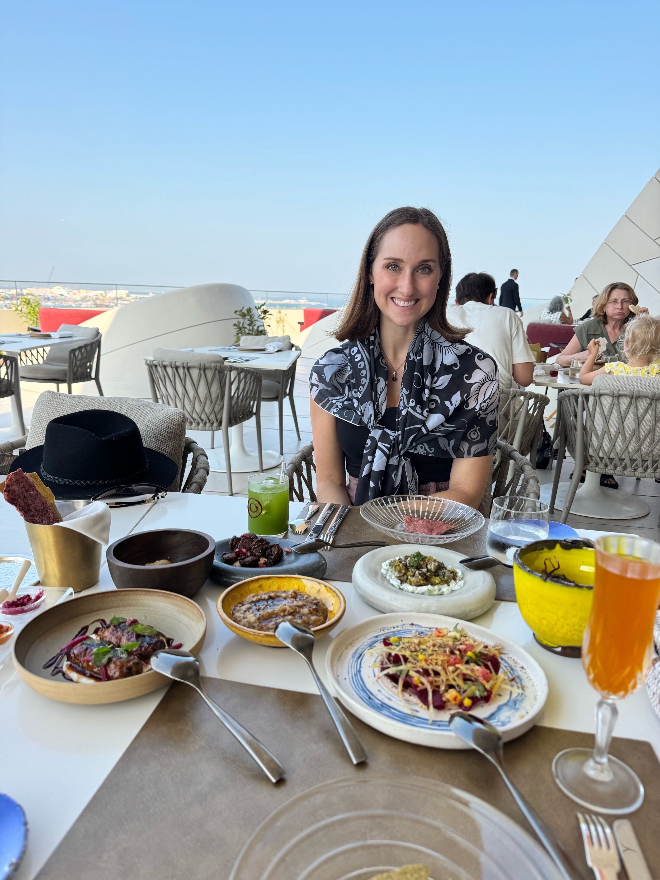 A woman smiling at a dining table outdoors with a city view in the background, surrounded by various plates of food and drinks.