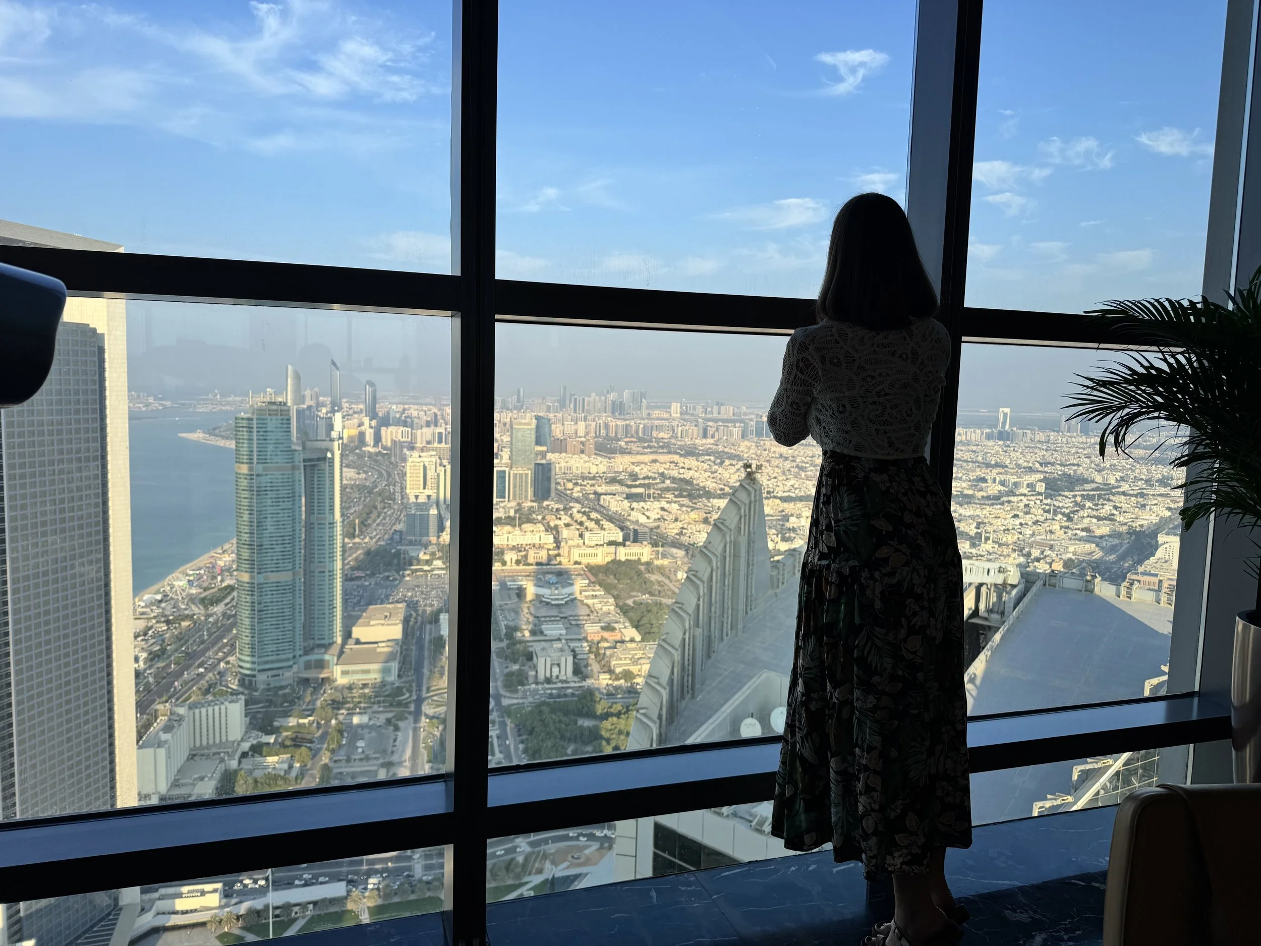 A woman standing near large floor-to-ceiling window, looking out over a cityscape with tall buildings and a river, during daytime.