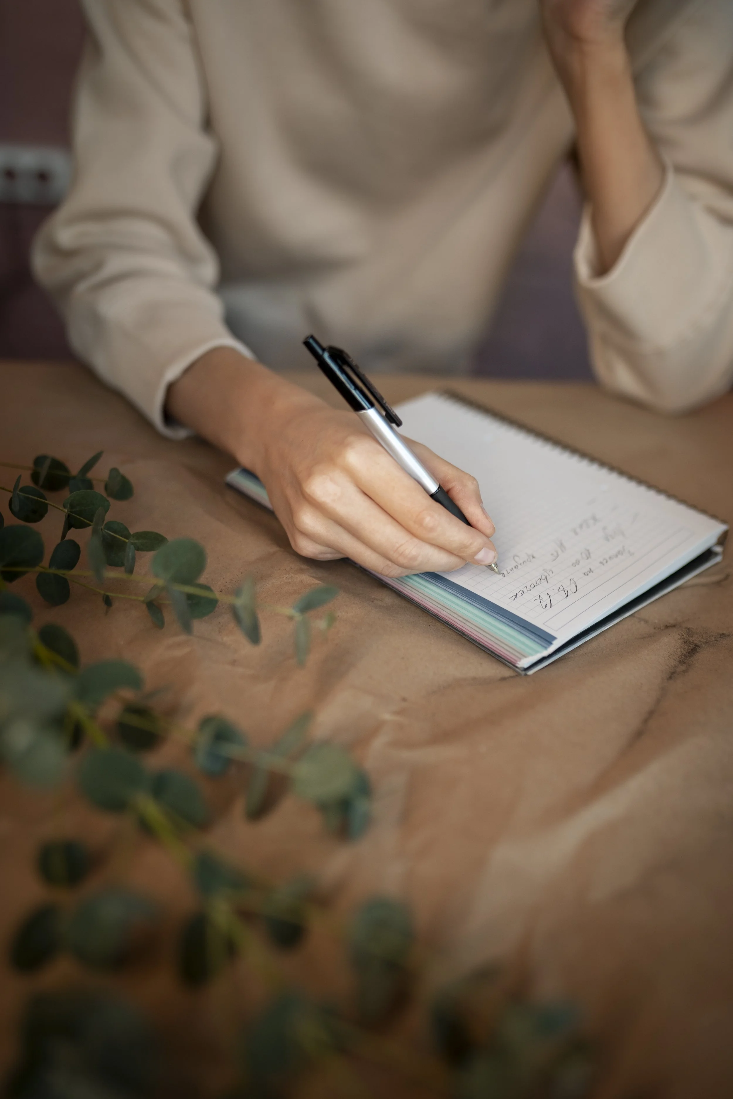 Person writing in a lined notebook with a black and silver pen, resting on a wooden table with a green plant in the foreground.