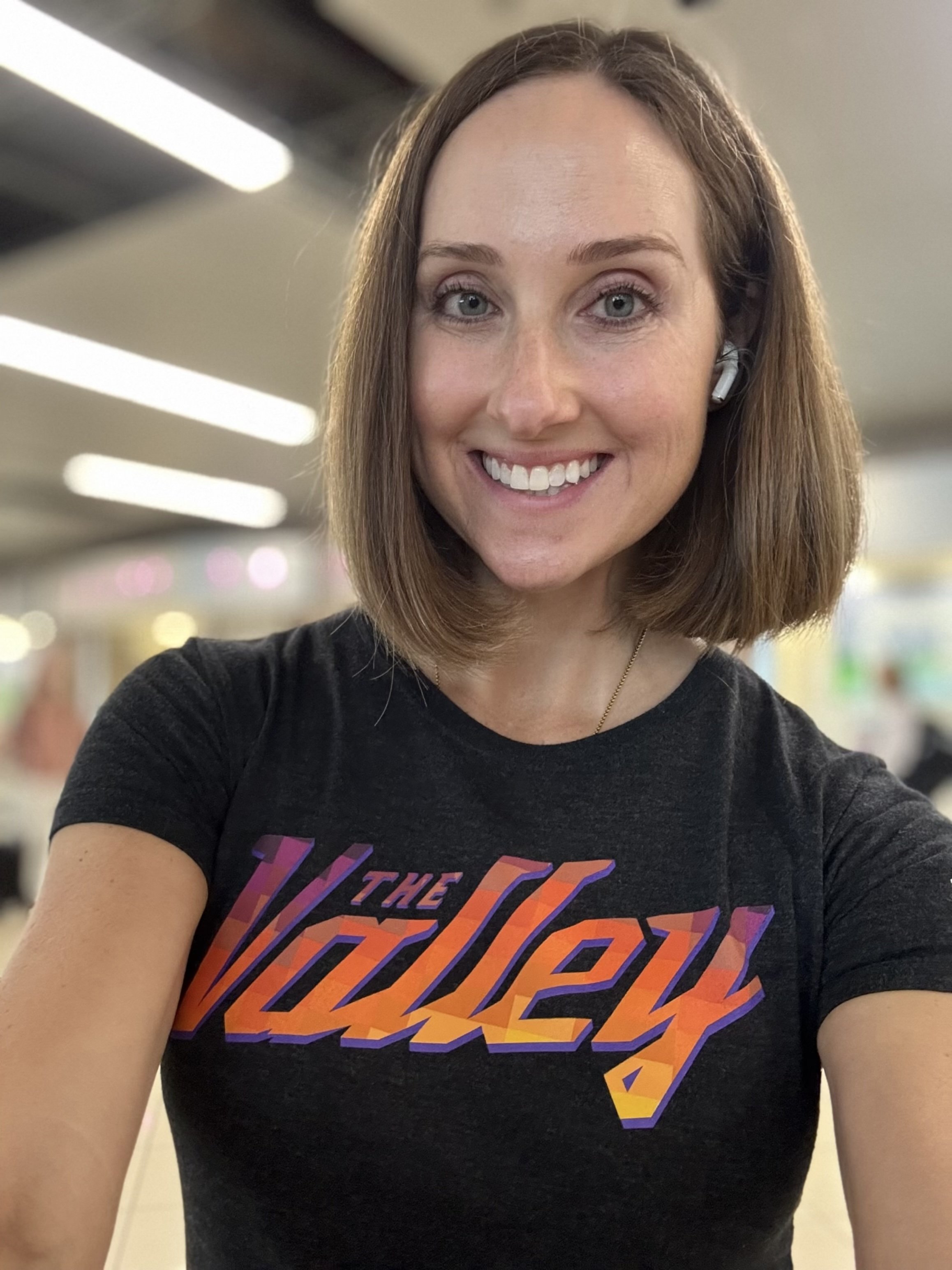 A woman with shoulder-length brown hair smiling and wearing a black t-shirt with colorful text that reads 'The Valley' in a brightly lit indoor environment.