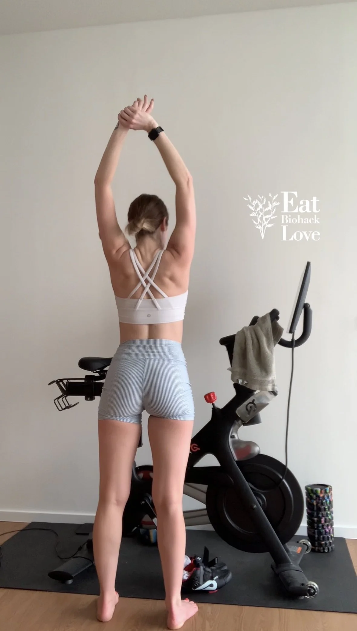 A woman standing in a room with her back facing the camera, stretching with her arms raised overhead, next to a stationary exercise bike. She is wearing a white sports bra and light blue shorts.