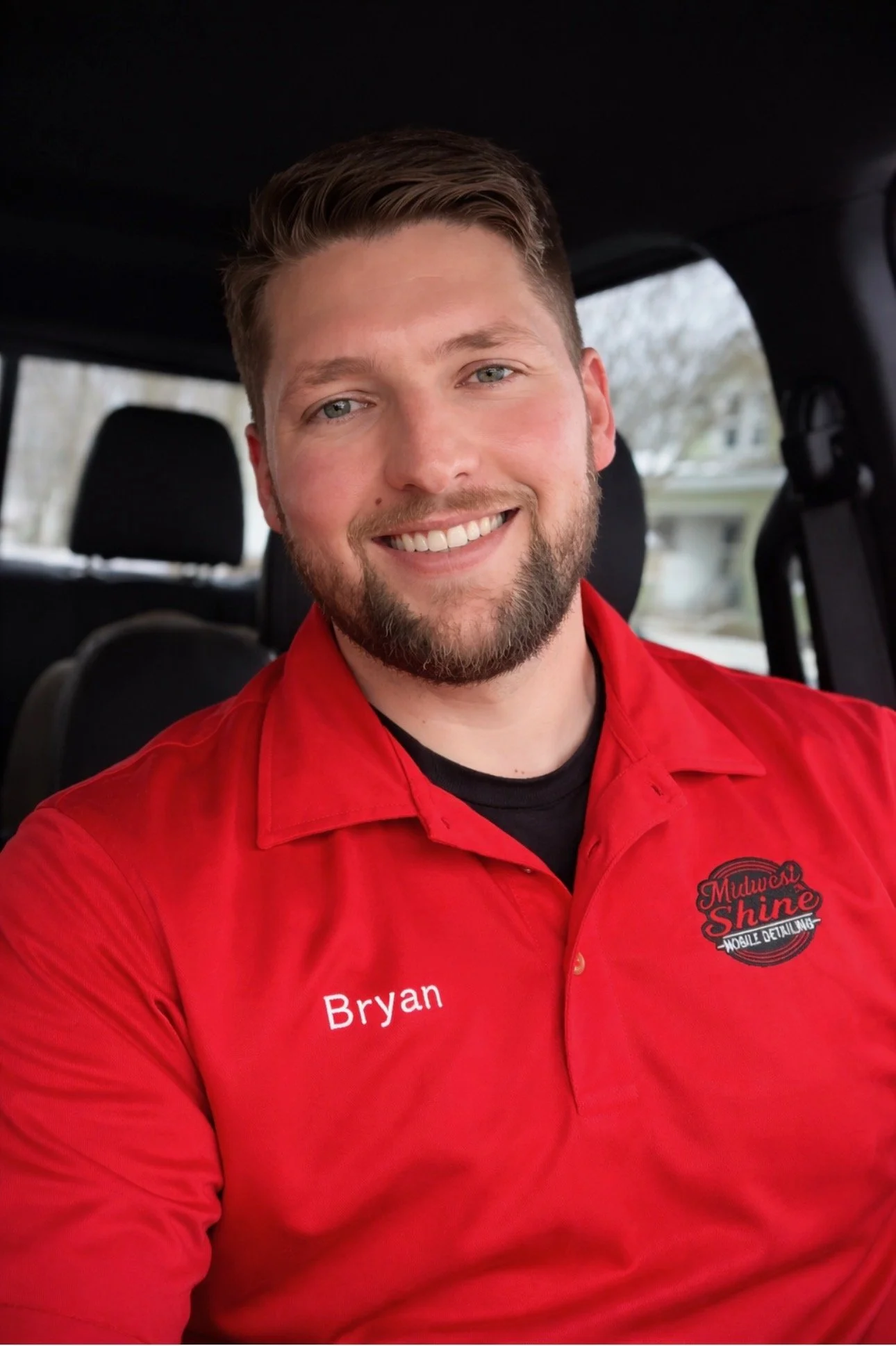 A smiling man with short brown hair and a beard, wearing a red shirt with "Bryan" embroidered on it, sitting inside a vehicle.