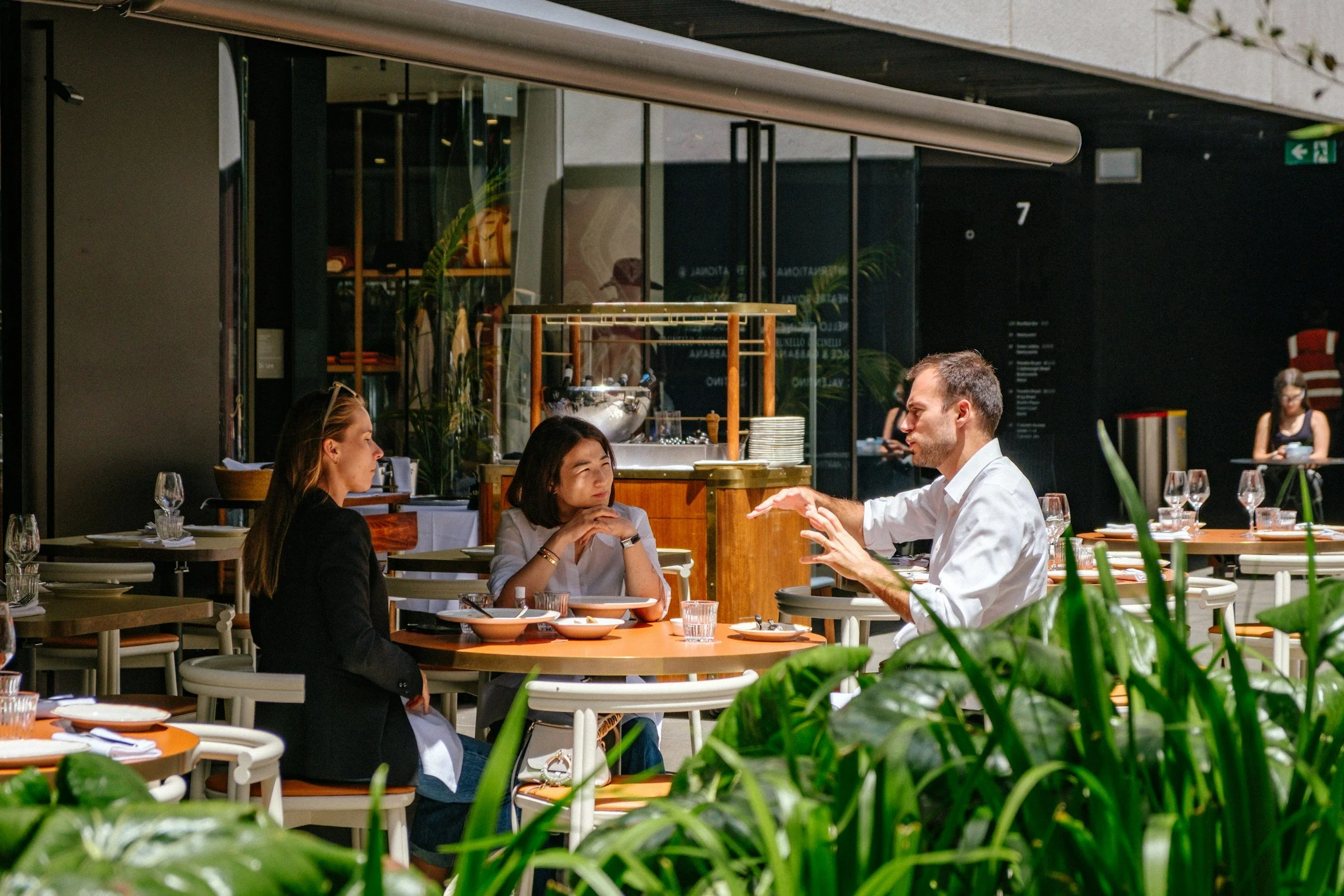 Four people in a restaurant, three seated at a table and one standing, engaging in conversation. The restaurant has large windows, plants, and table settings with glasses and plates.