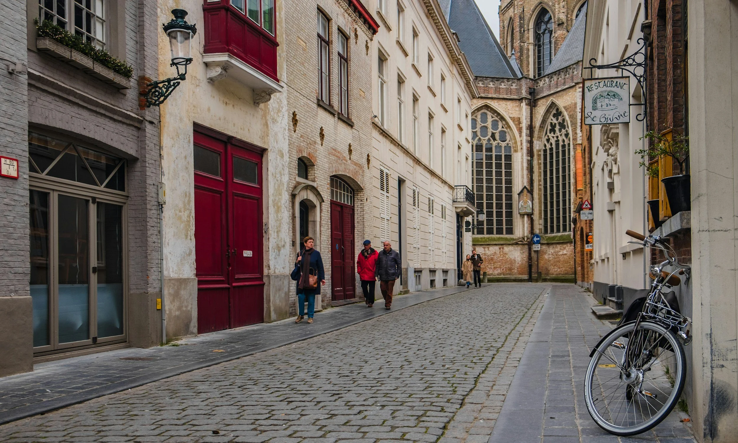 A cobblestone street in a European city with pedestrians, old buildings with red and white facades, and a parked bicycle, near a church with large stained glass windows.